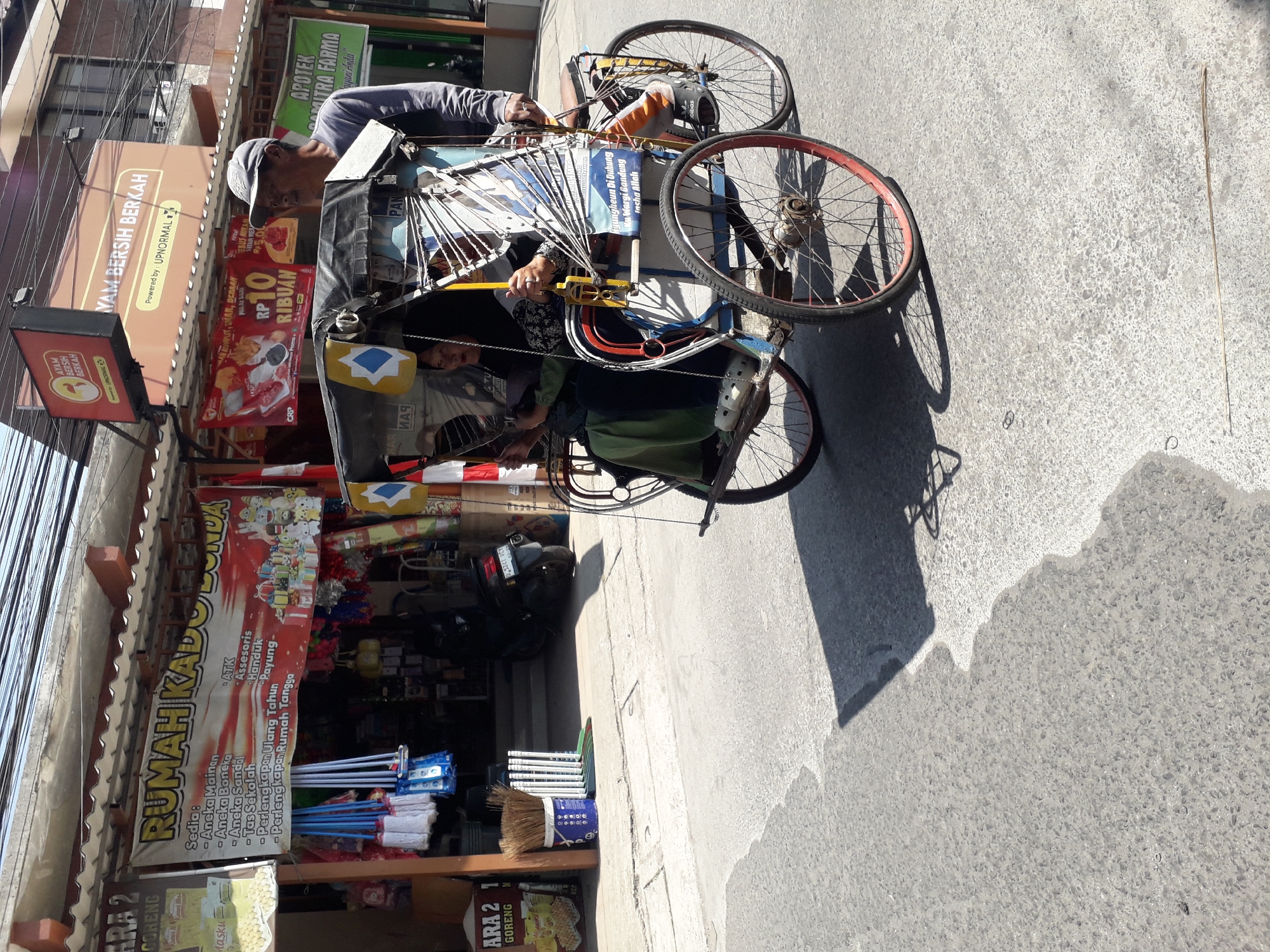 The image shows a cycle rickshaw traveling on a street in Linggar, Indonesia. The rickshaw, with a driver and two passengers, is partially covered by a black canopy. The driver, a man wearing a grey cap and long-sleeved shirt, is facing to the right. One passenger, a woman in a green headscarf and long green dress, is seated in the front, and another passenger, a woman in a black hijab and patterned dress, is seated behind her. The rickshaw has a metal frame, spoked wheels with red rims, and decorative flags. In the background, there are storefronts with various signs, including one that reads "RUMAH KADO LONDA" (Gift House LONDA) and another advertising "AYAM BERSIH BERKAH" (Clean Blessed Chicken). There are also parked motorcycles and hanging mops visible. The street is paved with asphalt, and shadows are cast from the buildings and the rickshaw. The lighting suggests it is daytime.
