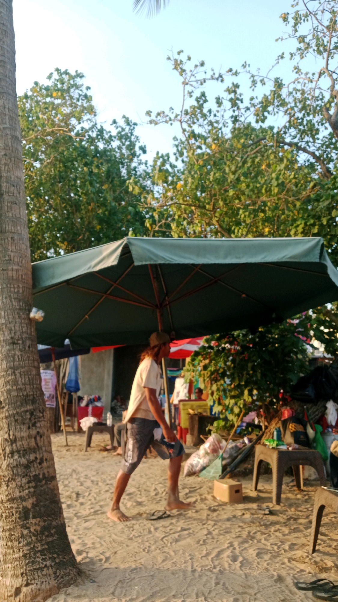 A man with long brown hair and a cap is walking on a sandy beach in Kuta, Indonesia, under a large green umbrella. He is wearing a light-colored t-shirt and patterned shorts. He is holding a plastic bag filled with items. The beach has scattered debris and a few small stalls set up with umbrellas. In the background, there are trees and a clear sky.