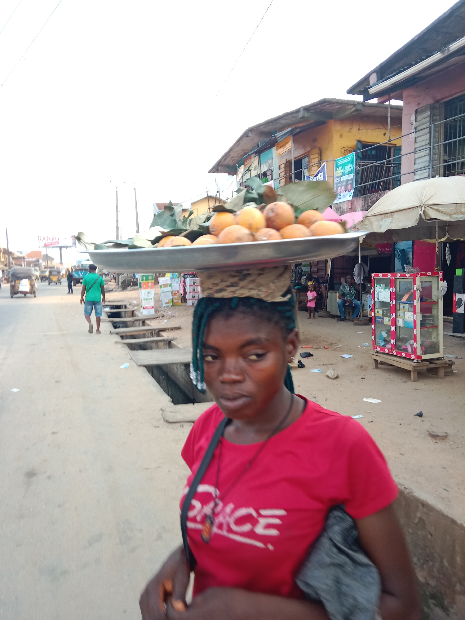 A young woman with blue braids walks on a partially paved street in Fegge, Nigeria, carrying a silver tray loaded with round, orange-colored fruits and green leaves on her head, supported by a woven pad. She wears a red t-shirt with white lettering and a dark shoulder bag. A shallow, open drainage ditch runs parallel to the street's edge. In the background, people walk along the street, and commercial buildings line the far side, including a yellow building with advertisements. A glass display cabinet stands near the buildings. Vehicles, including motorized tricycles, move in the distance.