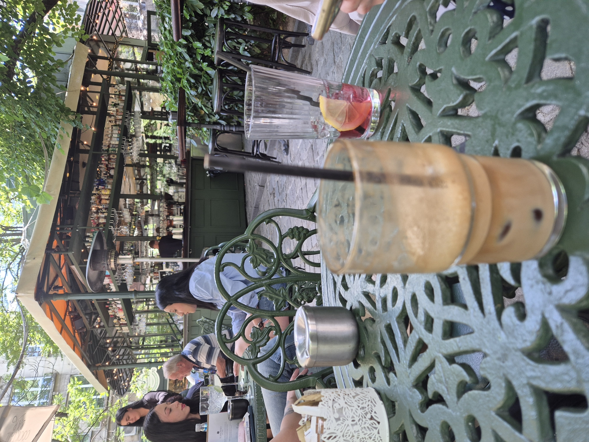 Outdoor cafe in Sofia, Bulgaria. A green, ornately patterned metal table is surrounded by patrons. One glass with a black straw holds a tan-colored liquid; another holds a red beverage with a lemon slice. An ashtray and a decorative tissue box sit on the table. People sit around the table, some with heads bowed and others looking into the distance. A bar is visible in the background, lined with bottles. Overhead, a retractable awning provides shade. Trees and buildings peek through the background.