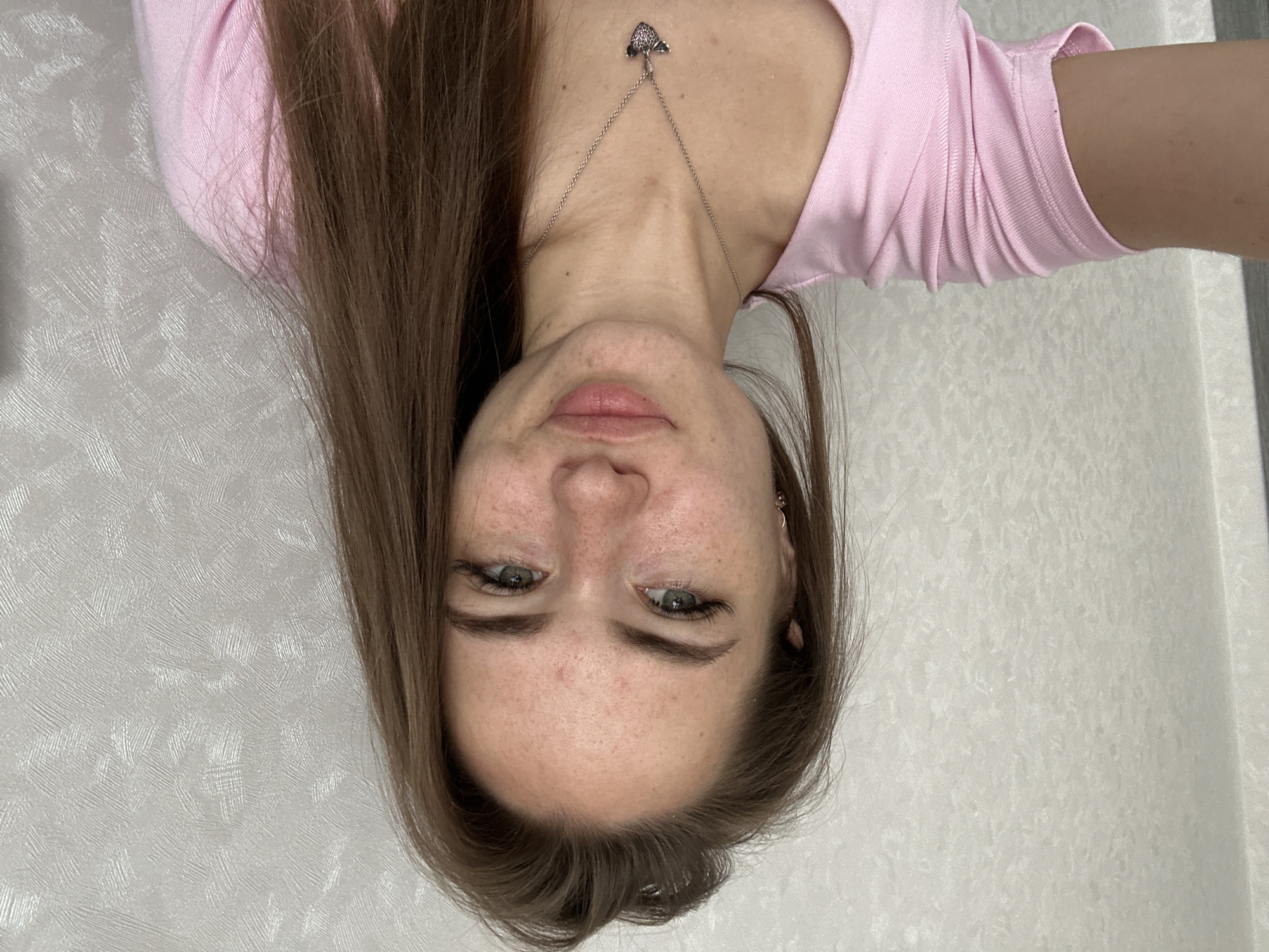 A young woman with long brown hair and green eyes smiles at the camera. She has freckles across her nose and cheeks and appears to have a few blemishes on her forehead. She is wearing a pink t-shirt and a silver necklace with a heart-shaped pendant. The background is a textured, light-colored wall. The image appears to be a selfie taken indoors.