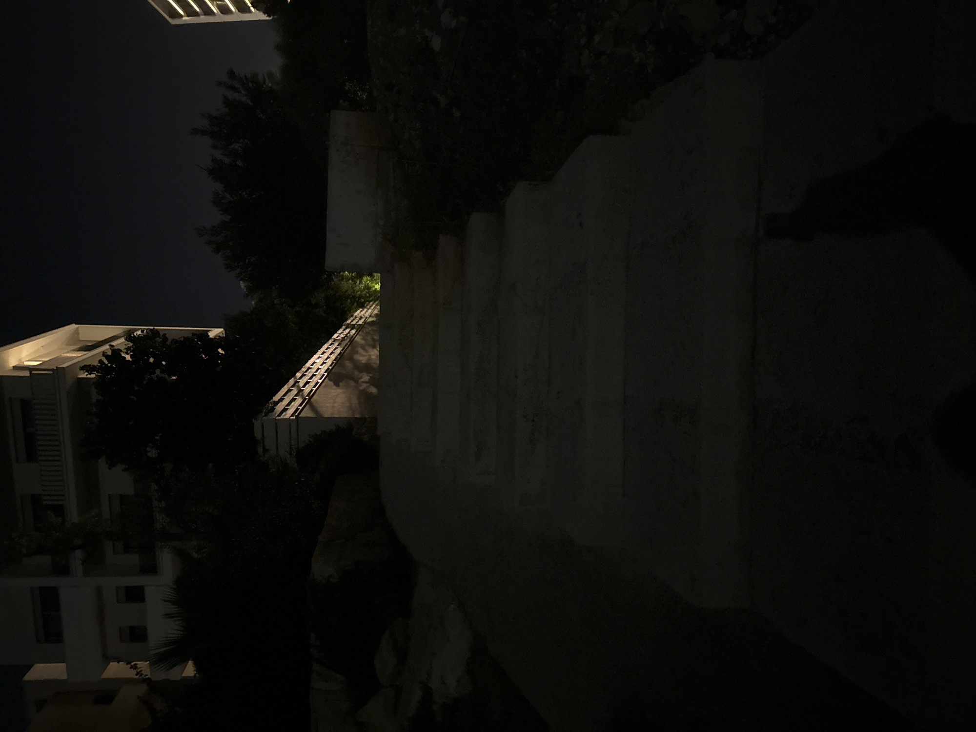 A night-time photograph shows a concrete staircase leading upwards. The staircase is dimly lit, with shadows cast on its surface. To the left of the staircase, there is a dark, rocky embankment with some vegetation. To the right, there is a white wall and more vegetation. In the background, partially visible, are buildings. One building appears to be a modern residential structure with balconies and windows. The sky is dark, indicating it is nighttime. The scene is located in Parekklisia Community, Cyprus.