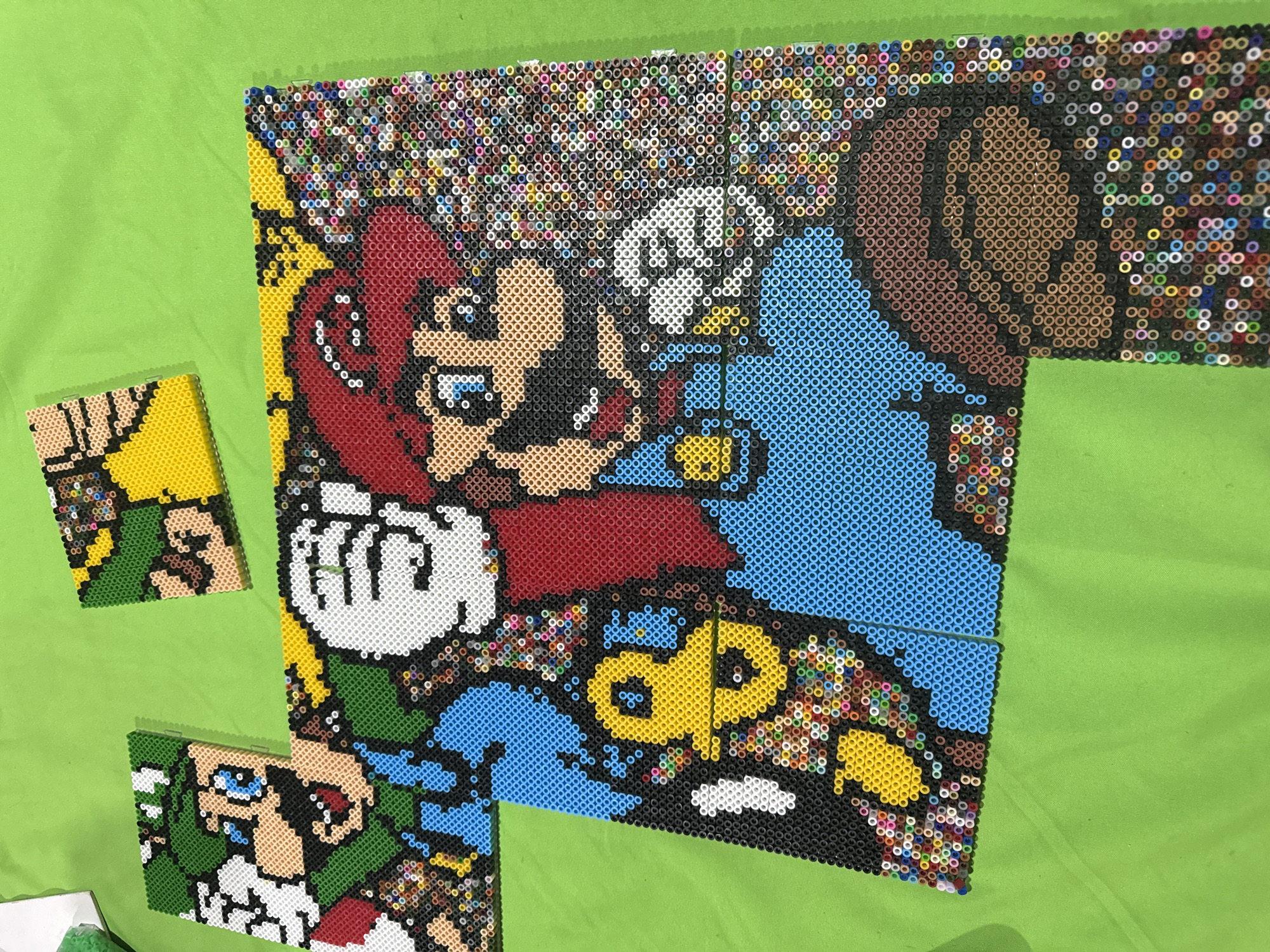 The image depicts a collection of pixel art pieces made from fused plastic beads, arranged on a light green fabric surface. The main piece is a large depiction of Mario and Donkey Kong, constructed from the beads. Two smaller, separate pixel art pieces depicting Link from the Legend of Zelda are also present. The location is not explicitly stated but can be pinpointed to Puebla, Mexico. Details about people and interactions are not present as the image focuses on the artwork itself. The environment is defined by the light green fabric backdrop.