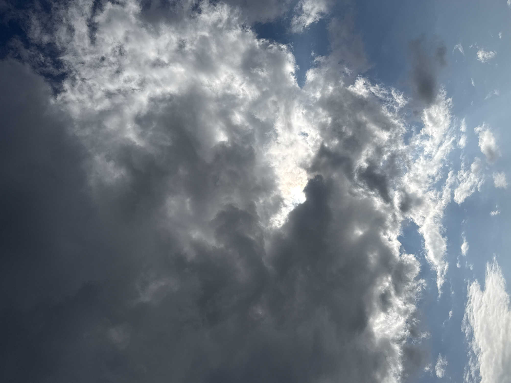 The image displays a dramatic sky filled with a mix of dark, heavy clouds and lighter, wispy ones. Sunlight peeks through gaps in the darker clouds, creating bright, illuminated patches. The blue sky is visible in the spaces between the clouds, especially towards the bottom and right of the frame. The overall impression is of a dynamic and possibly stormy sky. The image does not contain any people, subjects, or objects that would indicate a specific location within the city of Galinė, Lithuania.