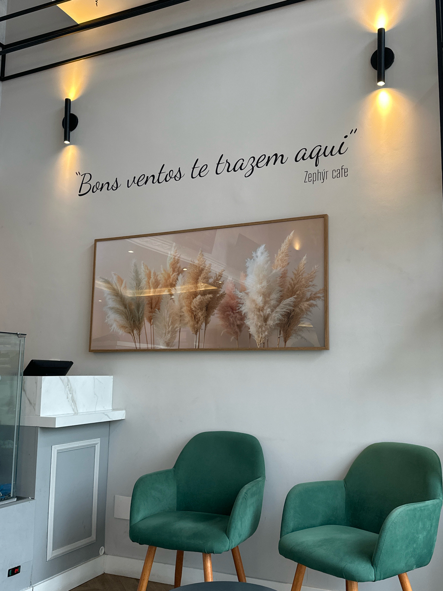 The image depicts an interior scene, likely a cafe or waiting area, with a focus on wall decor and seating. Two modern, black cylindrical wall sconces with warm light emanate from the upper left and right of the frame. Between them, on the white wall, is a black cursive inscription that reads, "Bons ventos te trazem aqui" followed by "Zephyr cafe" in a smaller font. Below the inscription, a framed rectangular artwork displays a collection of pampas grass plumes in warm, neutral tones against a soft pink background. To the left of the artwork, a sleek black tablet-like device rests on a white marble-topped counter. The counter is supported by a grey cabinet with a decorative white trim. In the foreground, two plush, emerald green armchairs with wooden legs are positioned facing each other. The overall ambiance suggests a stylish and inviting space. The specific location of Florianópolis, Brazil, is not discernible from the visual information provided in the image.
