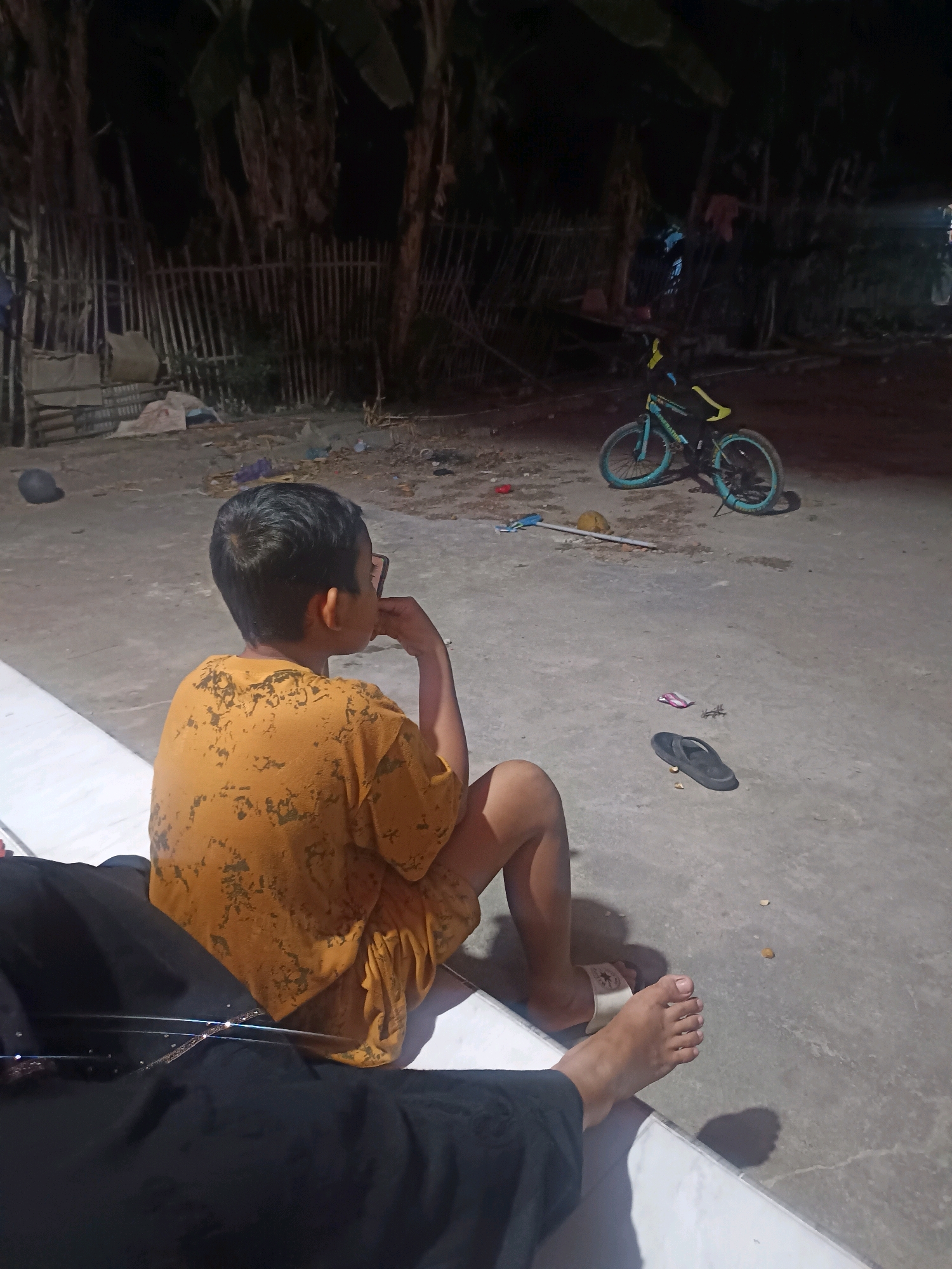 In Gunung Sekar, Indonesia, a young boy wearing a yellow t-shirt with a textured print sits on a concrete ground at night. He is facing away from the camera, with his right elbow resting on his knee and his chin in his hand, looking towards the right. His left leg is bent, and his right leg is extended. Beside him, partially visible, are the legs of another person dressed in black. A blue and green bicycle with yellow handlebars is parked in the background. Scattered on the ground are various items including a pair of black flip-flops, a small pink object, and debris. The environment appears to be an outdoor area with a fence and trees in the background, dimly lit by artificial light.