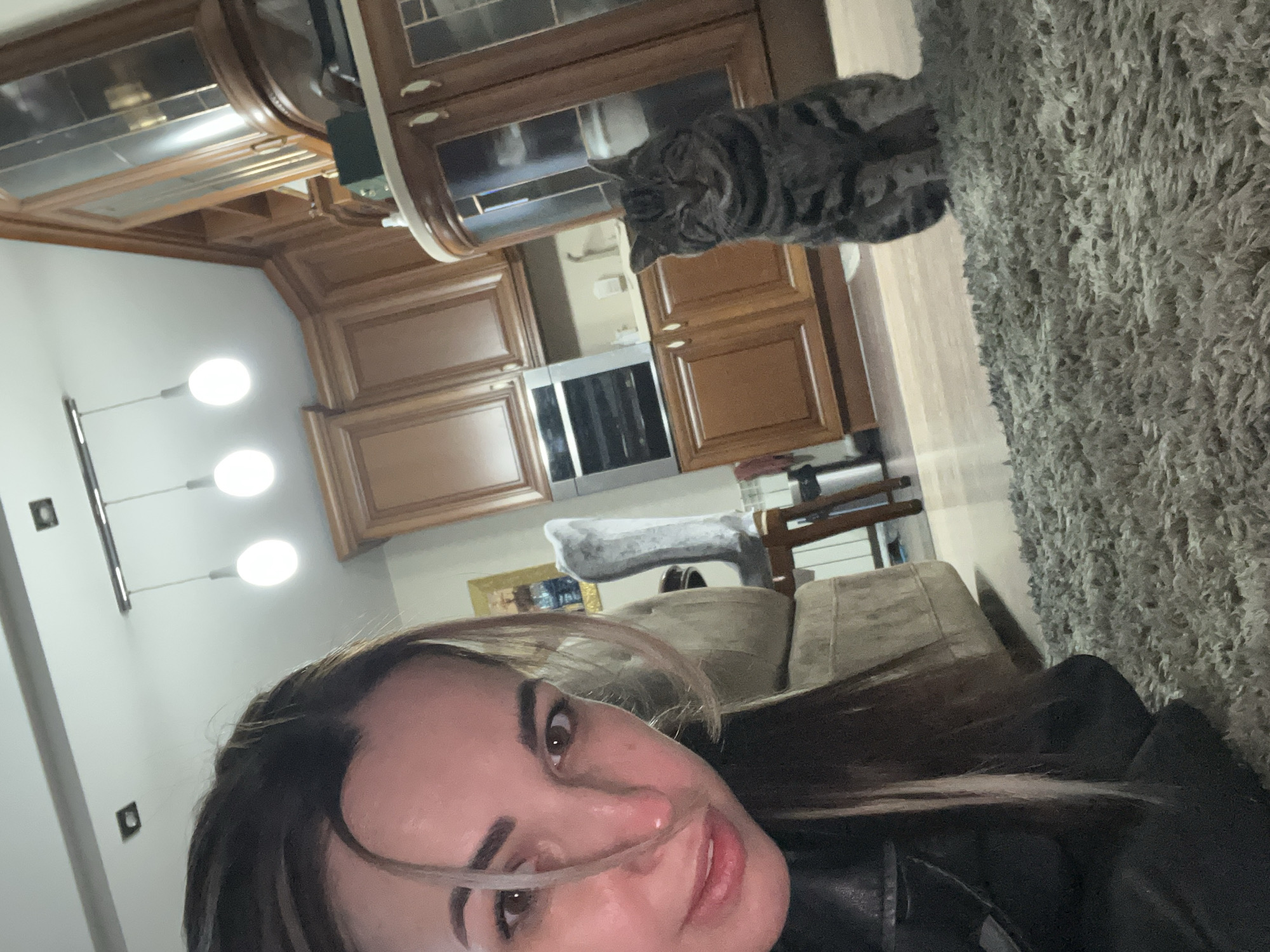 A woman is taking a selfie in a kitchen. A grey tabby cat is on top of a kitchen cabinet. The kitchen features wooden cabinets, a silver oven, and a modern lighting fixture. A grey sofa and rug are in the foreground. The location is Unknown, Unknown.