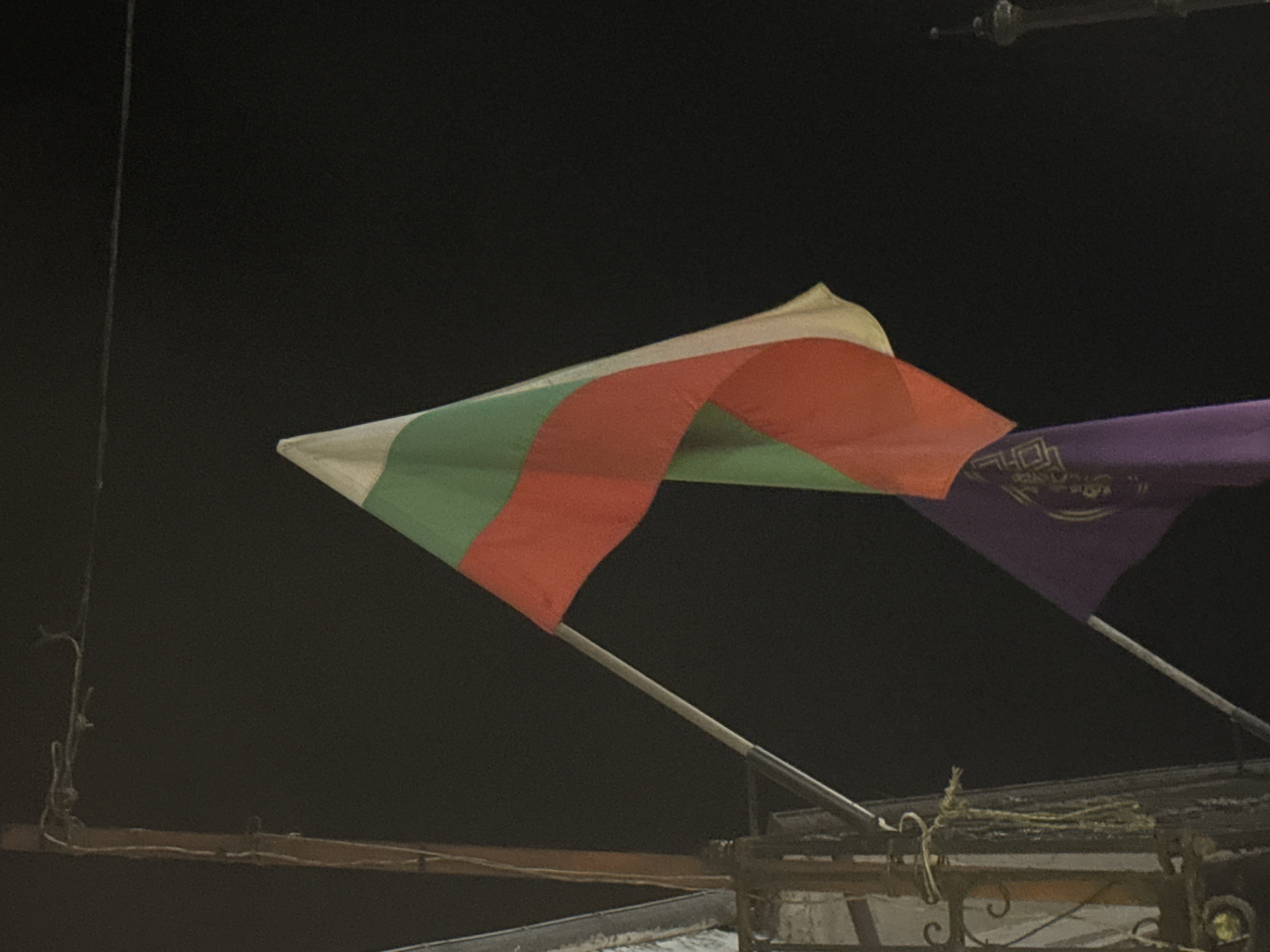 A night shot depicts two flags waving atop a building or structure in Veliko Tarnovo, Bulgaria. The flag on the left is the Bulgarian national flag, with horizontal white, green, and red stripes. To the right is a purple flag bearing illegible gold text or symbols. The flags are mounted on metal poles. Below, there is a metal structure and what appears to be a roof. The background is a dark night sky.