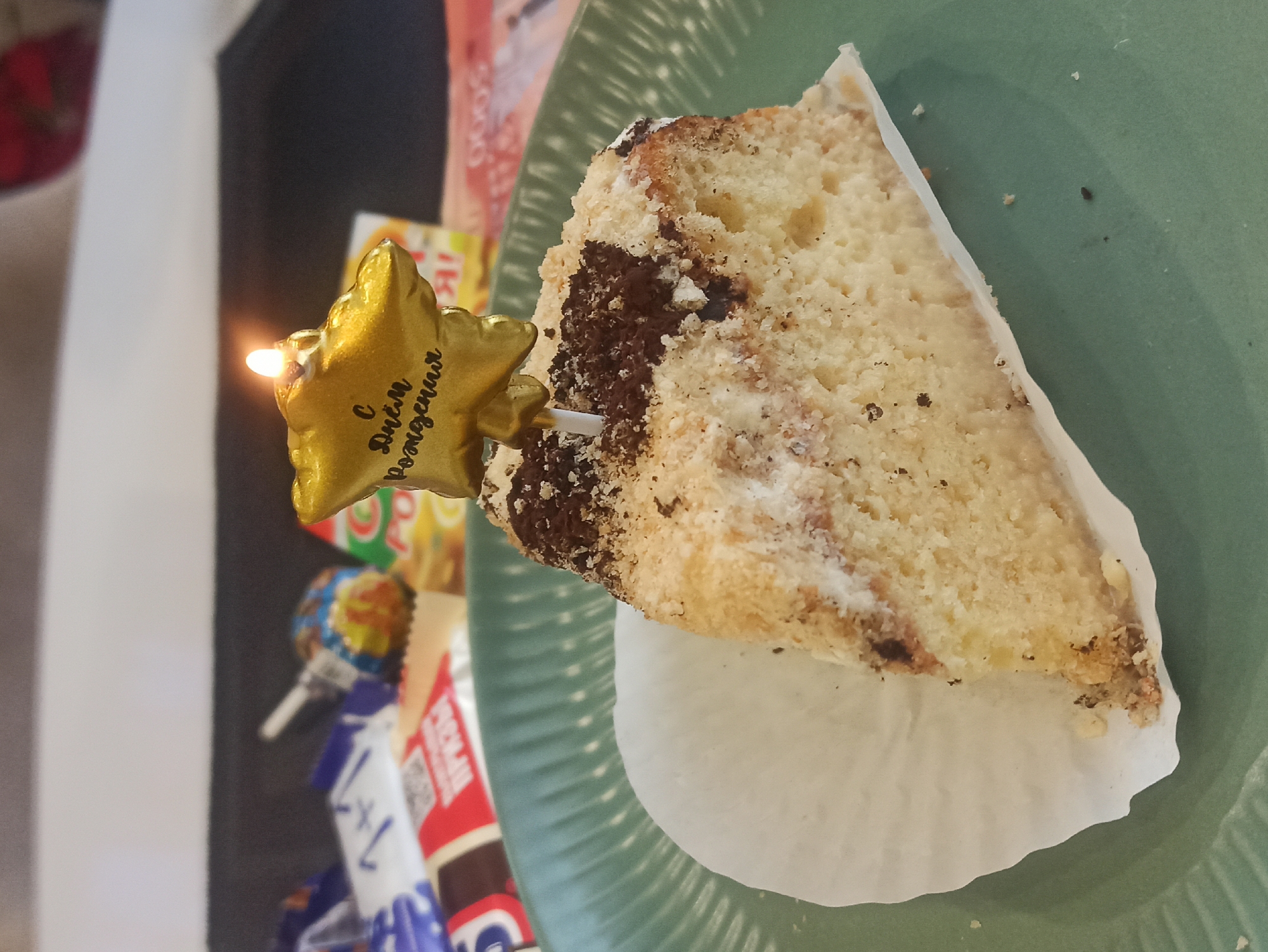 A slice of cake rests on a paper doily, placed on a green plate. A lit candle shaped like a golden star, inscribed with Cyrillic characters (С Днём Рождения), is stuck into the cake. Candies and other food items are visible behind the plate. The location is Кондратово, Russia.