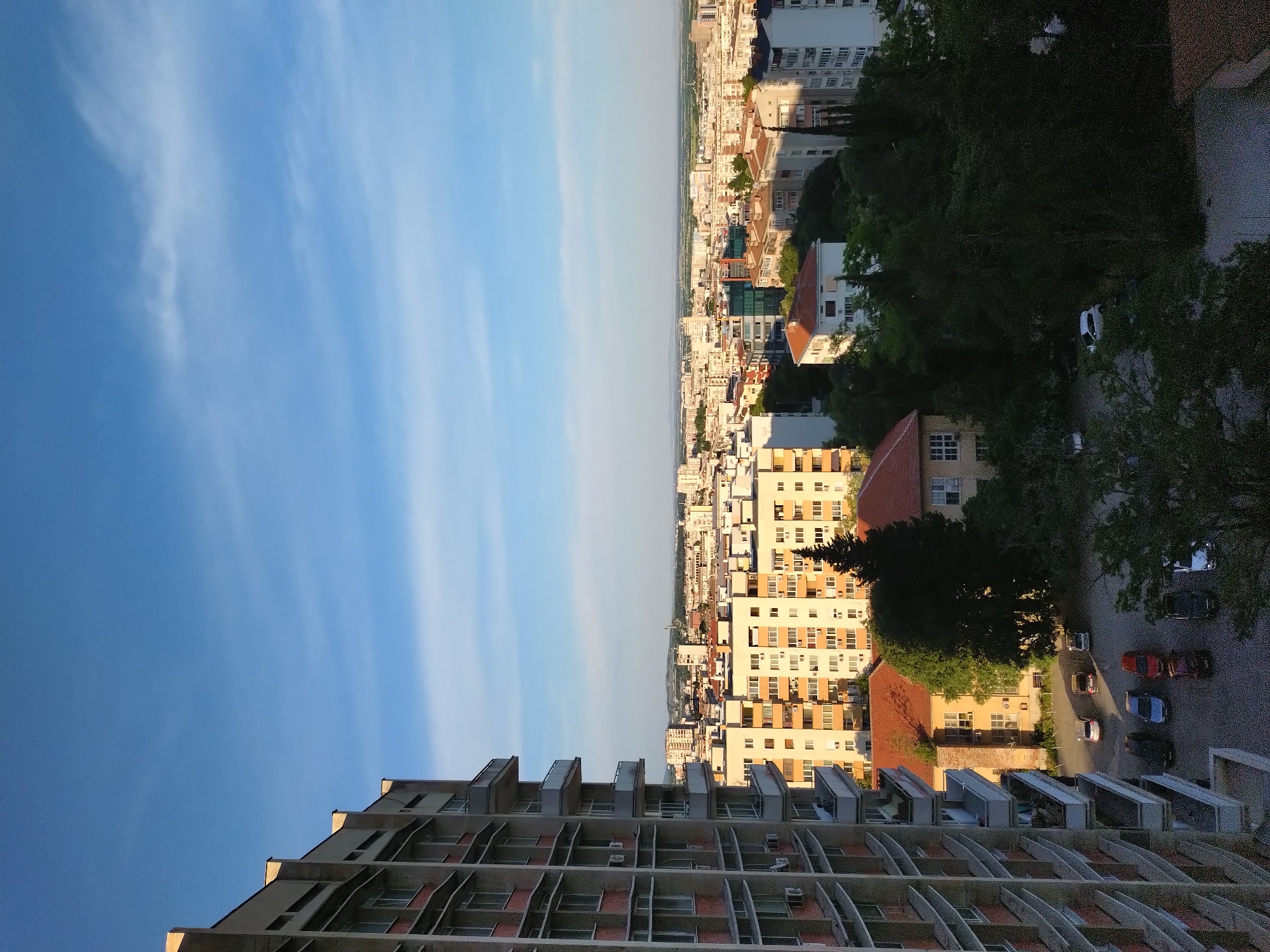 This is a high-angle, eye-level, outdoor, vertical shot of a city skyline in Stara Zagora, Bulgaria, during the day. The foreground is dominated by the side of a large, multi-story apartment building with visible balconies and windows. The midground features a dense collection of other apartment buildings and houses, some with red tile roofs, interspersed with trees. A parking area with several cars is visible in the lower midground. The background shows a further expanse of urban development that fades into a hazy horizon. The sky is a clear blue with thin, wispy clouds. The scene is bathed in warm sunlight, casting shadows and highlighting the architectural details of the buildings. There are no visible people or animals.