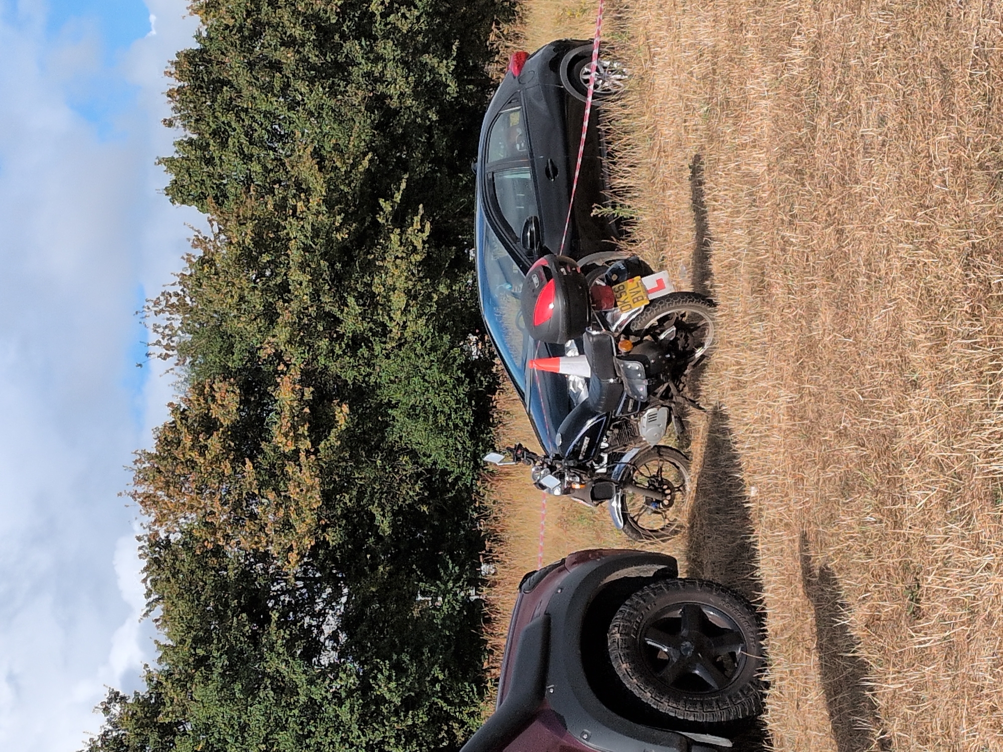 An outdoor scene features a dry, grassy field with a line of trees in the background. In the foreground, a portion of a dark-colored SUV is visible on the left, with a black motorcycle parked in the center. A black car is parked to the right of the motorcycle. A red and white striped tape runs across the scene, separating the motorcycle and car from the foreground. A traffic cone is placed on the motorcycle's rear luggage. The sky is cloudy. The location appears to be a rural area, possibly in or near South Staffordshire, United Kingdom.