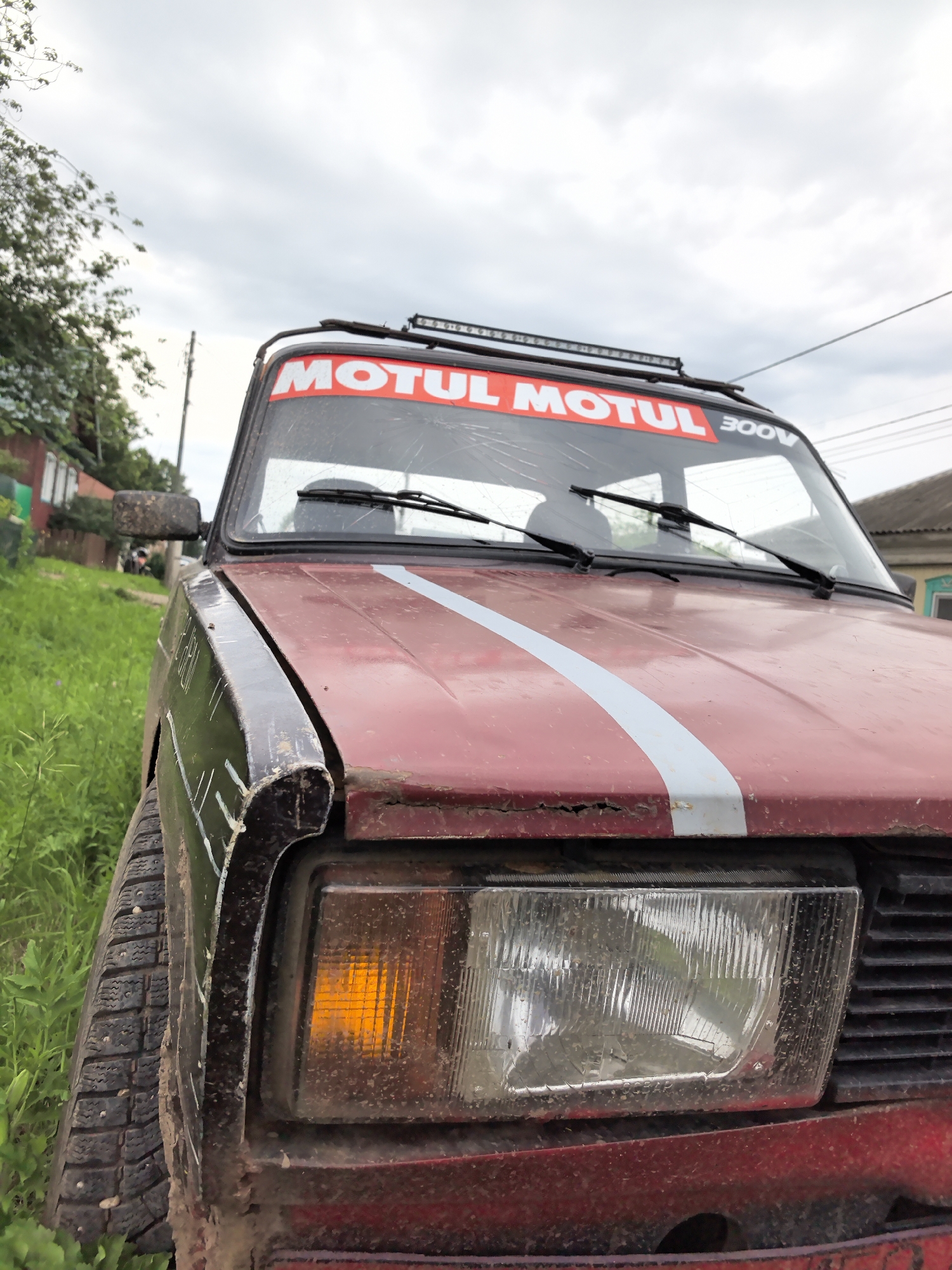 A close-up, low-angle exterior shot of the front of a red car shows a muddy, worn condition. The car is parked outdoors, next to a grassy area and a fence, suggesting a rural or suburban setting, possibly in the village of Koyanovo, Russia, given the context. The car's windshield has a red banner with white lettering that reads "MOTUL" and "300V." The windshield is cracked. A light bar is mounted on the roof of the car. The right front headlight is illuminated with an orange tint, and the left headlight is visible but appears dull. The front fender is dark colored and appears damaged. The car's tire is visible on the left side of the frame, covered in mud. In the background, there is a house with a gray roof and some trees. The sky is overcast.