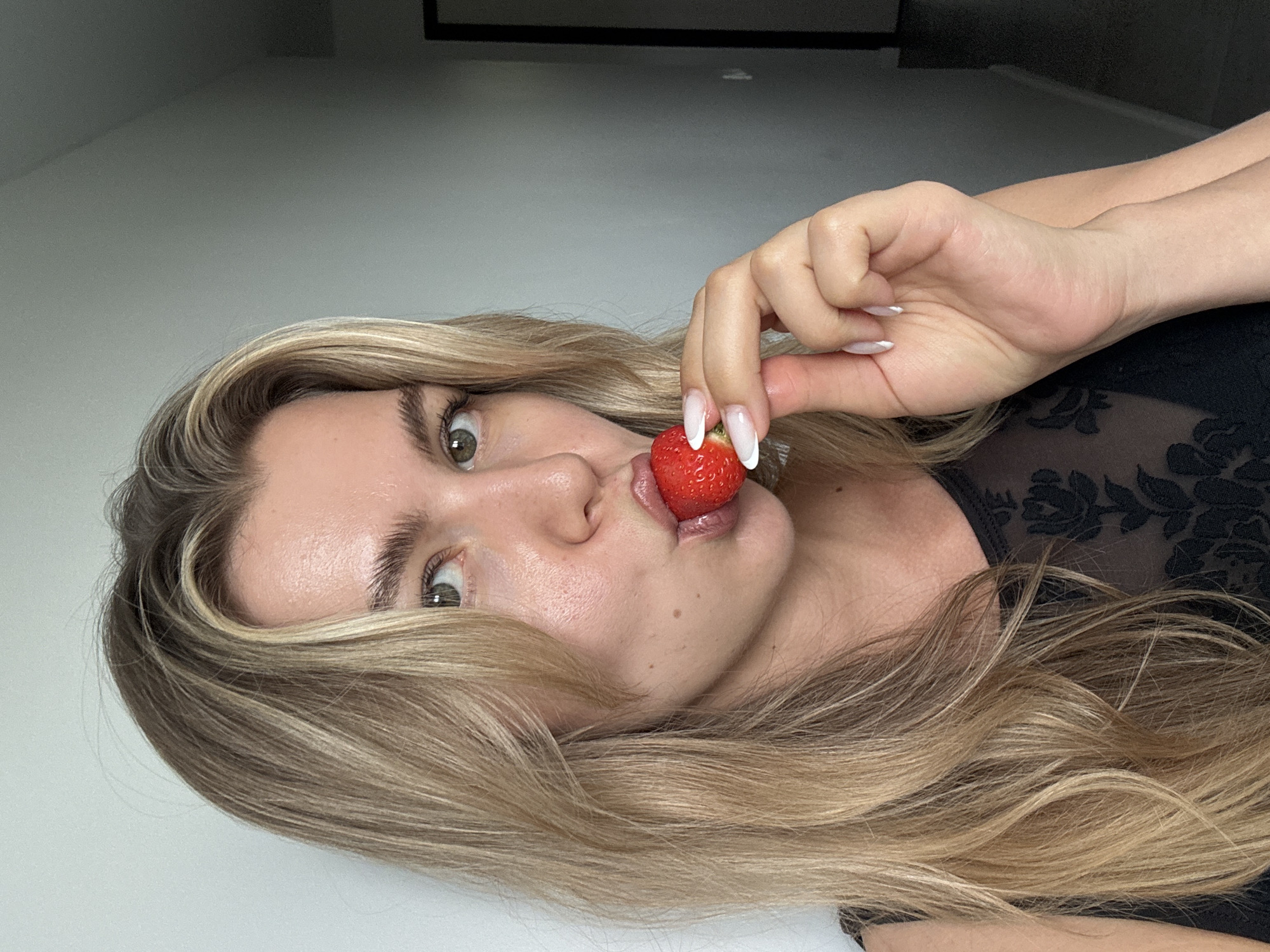 A medium close-up shot shows a young Caucasian woman with blonde hair and green eyes holding a strawberry to her lips. She is wearing a black top with a lace floral pattern. The background is a plain wall.