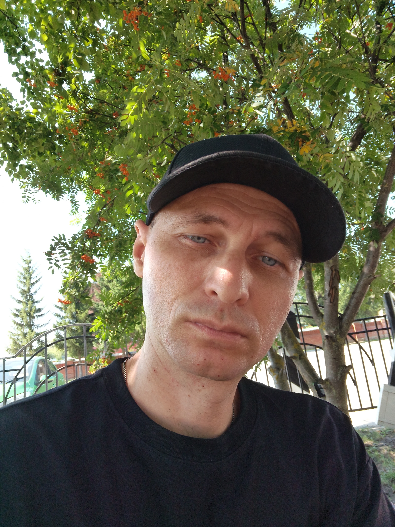 The image is a close-up, over-the-shoulder selfie taken outdoors, likely in Mokshan, Russia, given the context. A man wearing a black baseball cap and a black t-shirt is the primary subject. He has light-colored skin, blue eyes, and is looking towards the camera with a neutral expression. The background features a tree with green leaves and small orange berries, suggesting it might be late summer or early autumn. Partially visible in the background is a metal fence, a portion of a light green car, and a tall evergreen tree. The lighting indicates it is daytime, possibly sunny, with some shadows cast by the tree.
