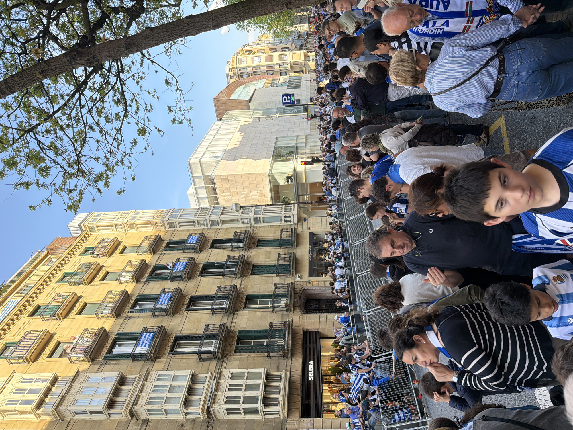 The image depicts a vibrant street scene in San Sebastián, Spain, under a clear, sunny sky, capturing a large crowd gathered for a public event. The photograph is oriented vertically, showcasing a street lined with multi-story buildings of diverse architectural styles.
A multitude of people, encompassing adults, teenagers, and children, are amassed along the street, positioned behind metal crowd control barriers. Many individuals are conspicuously dressed in blue and white striped attire, including shirts, scarves, and hats, signifying their support for a sports team. Notably, a scarf worn by an older man bears the text "AURRERA REALA," indicating allegiance to the local football club, Real Sociedad. The crowd's attention is primarily directed towards the street, suggesting they are observing a parade or a similar procession.
The buildings flanking the street include an older, ornate structure with numerous balconies, many of which are decorated with blue and white striped flags or banners, further emphasizing the team spirit. Adjacent to these are more contemporary buildings with glass facades. Trees with green foliage line the thoroughfare, hinting at a spring or summer season.
Visible text includes "SELERA" on a shopfront at street level, and a blue parking sign with a white 'P' accompanied by the text "20 MINUTU HOBETO" (20 minutes better). A red traffic light is also discernible. The overall atmosphere appears festive and engaged, illuminated by the bright daylight.
