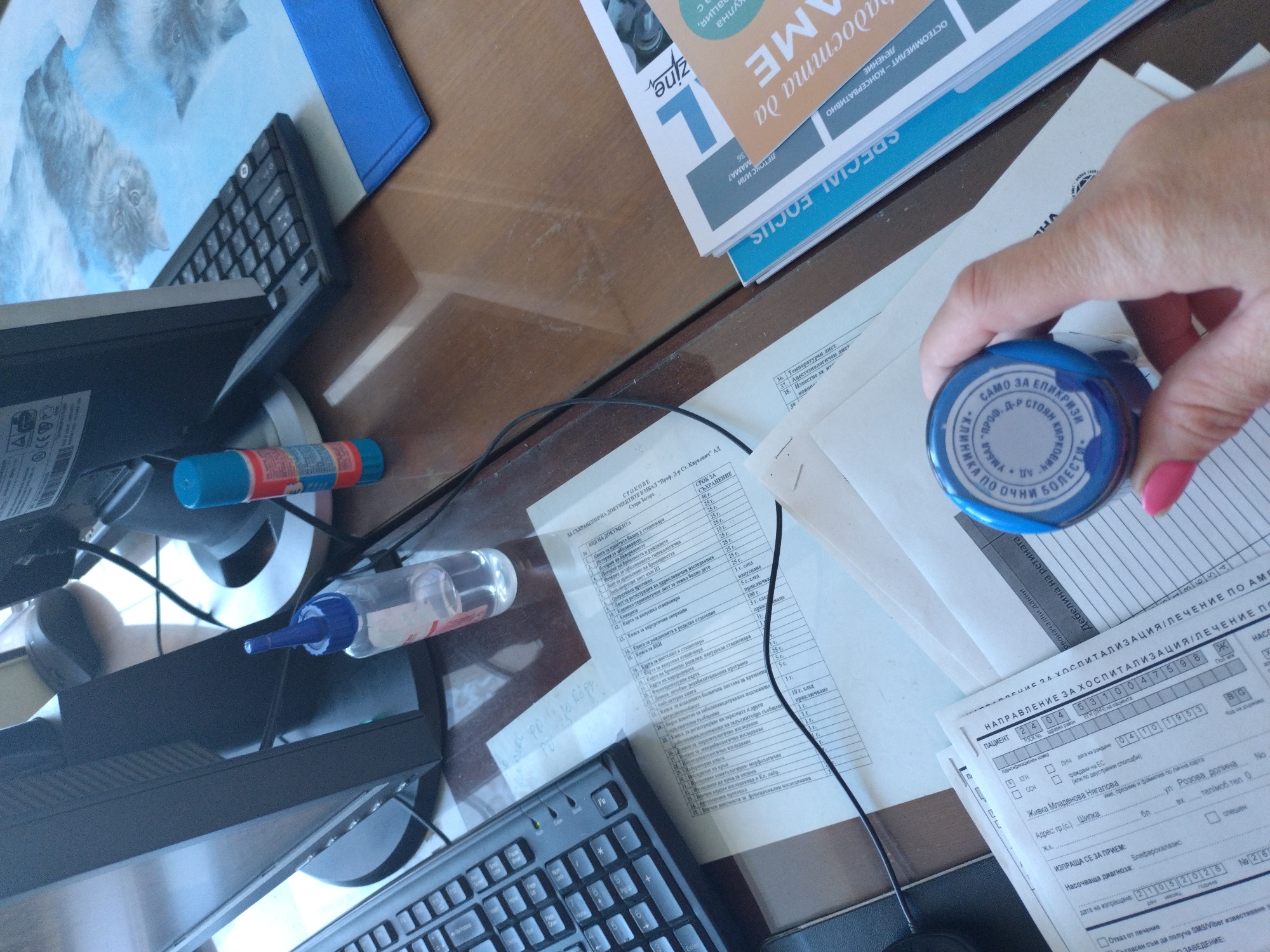 The image depicts a desk cluttered with office supplies and paperwork. In the center, a hand holds a blue rubber stamp with red ink, inscribed in Cyrillic, seemingly ready to stamp a medical form or document. The form is titled "Направление за хоспитализация/Лечение по АМЕ" (Referral for Hospitalization/Treatment under AME), and includes personal details like name, date of birth, address, and diagnosis. Scattered around are a computer monitor and keyboard, a glue stick, a bottle of liquid correction fluid, a magazine with a "SPECIAL FOCUS" banner, and other documents, including one titled "СРОКОВЕ ЗА СЪХРАНЕНИЕ НА ДОКУМЕНТИТЕ В МБАЛ 'Проф. Д-р Ст. Киркович' АД Стара Загора" (Document Retention Periods at MBAL 'Prof. Dr. St. Kirkovich' AD Stara Zagora), indicating the scene's location. A picture of cats on a blue folder is also visible in the upper left corner.