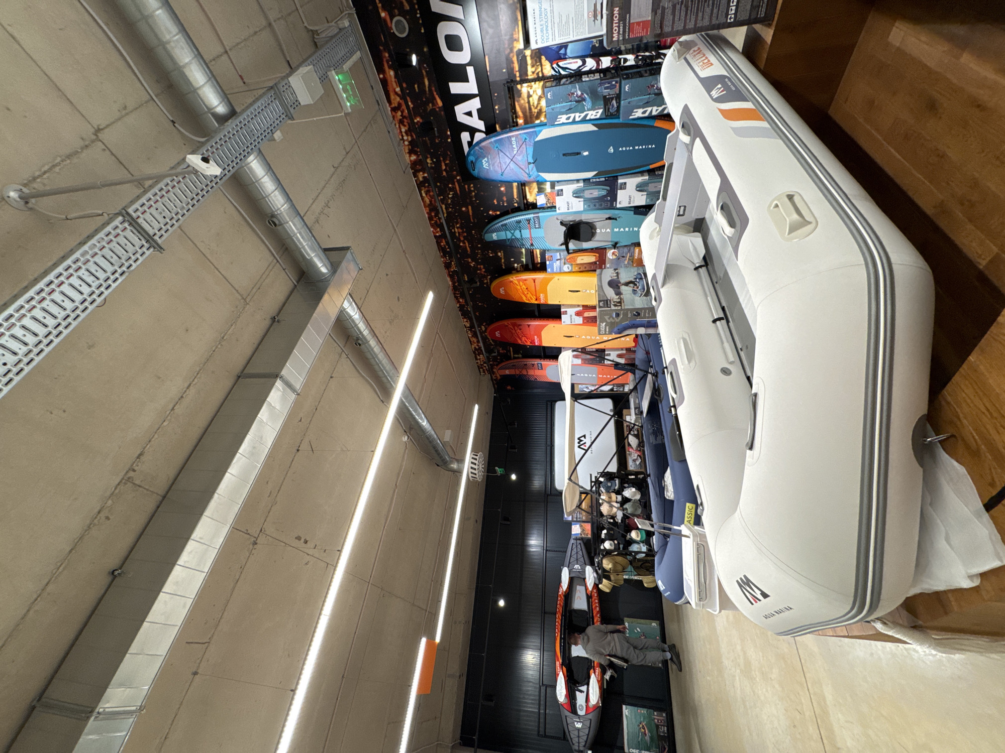This media file captures the interior of a large retail store, likely a sporting goods or outdoor equipment shop, located in Sofia, Bulgaria. The image is oriented vertically but depicts a wide perspective of the store's display area.
The primary subjects are water sports equipment. In the foreground and right side, a large, white inflatable boat, specifically an "Aqua Marina Deluxe" model, is prominently displayed on a wooden floor section. It features grey and orange accents and has "AQUA MARINA" branding visible. Behind it, other inflatable boats and kayaks are arranged. A distinctive orange and black kayak, with "WV" branding, is displayed on a black platform, with a person in a grey outfit standing beside it, partially obscured, appearing to interact with the display.
The background features a large black wall banner with water-themed imagery and various stand-up paddleboards (SUPs) displayed vertically. Models such as "AQUA MARINA BLADE," "AQUA MARINA BEAST," and "AQUA MARINA CLASSIC" are clearly visible, showcasing different designs and colors. Display information next to the SUPs includes phrases like "DOUBLE STRINGER TECHNOLOGY" and "MOTION." The brand "SALON" is partially visible at the top of the banner.
The store's ceiling is high and features an industrial aesthetic with exposed concrete, extensive metallic ventilation ducts, and cable trays running across. Bright, linear LED strip lights are installed along the ceiling, providing ample illumination. A green "EXIT" sign is visible on one of the ceiling structures. The overall scene suggests a well-lit, spacious retail environment dedicated to showcasing water recreation gear.