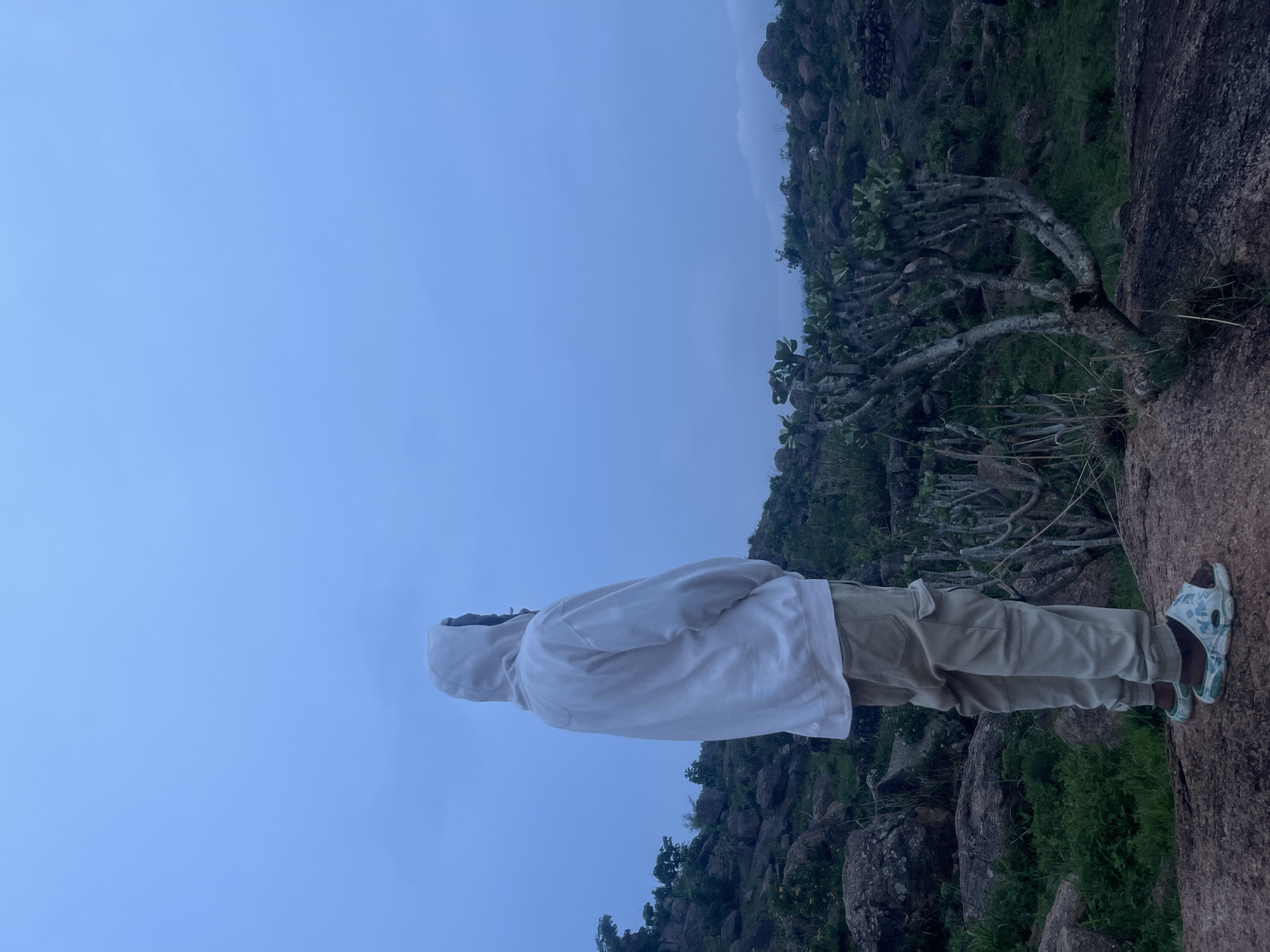 A person stands on a rocky outcrop, facing away from the camera and looking out over a landscape. The person is wearing a light-colored hooded sweatshirt and light-colored cargo pants, with patterned sandals on their feet. The environment is characterized by large rocks, sparse vegetation, and a cloudy sky. The scene appears to be outdoors in a somewhat rugged, natural setting, consistent with areas found in the city of Jos, Nigeria, particularly in its rocky terrain and open landscapes. The lighting suggests it is either early morning or late evening.