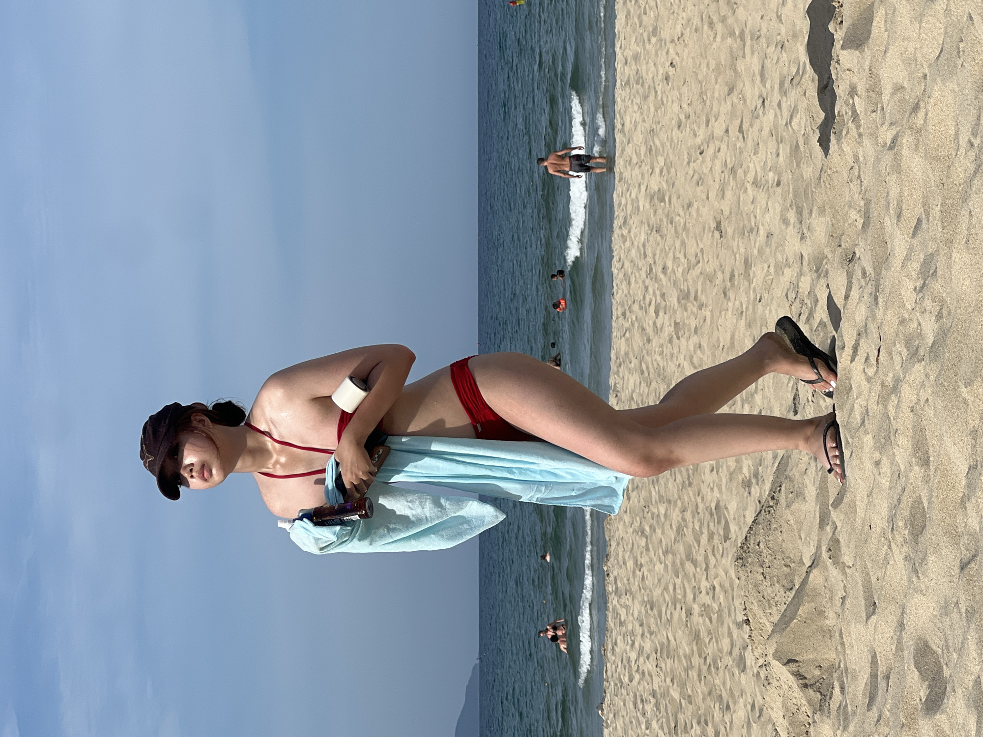 A young woman wearing a red bikini and a brown baseball cap walks on a sandy beach in Đà Nẵng, Vietnam. She holds a light blue towel draped over her left arm, and in her right hand, she carries a bottle of sunscreen and a white cylindrical object, possibly a lotion bottle or speaker. She is looking upwards and to her right, her skin glistening with moisture. Her feet are adorned with black flip-flops. The beach is mostly empty, with a few other people visible in the shallow water, some swimming and others standing. A man in dark shorts is wading in the ocean in the background, facing away from the camera. The sea is calm with small waves, and the sky is clear blue. In the far distance, a hazy coastline is visible. The sand is light brown and textured, with a small pile of sand to the left of the woman. The scene captures a moment of leisure on a sunny day at the beach.