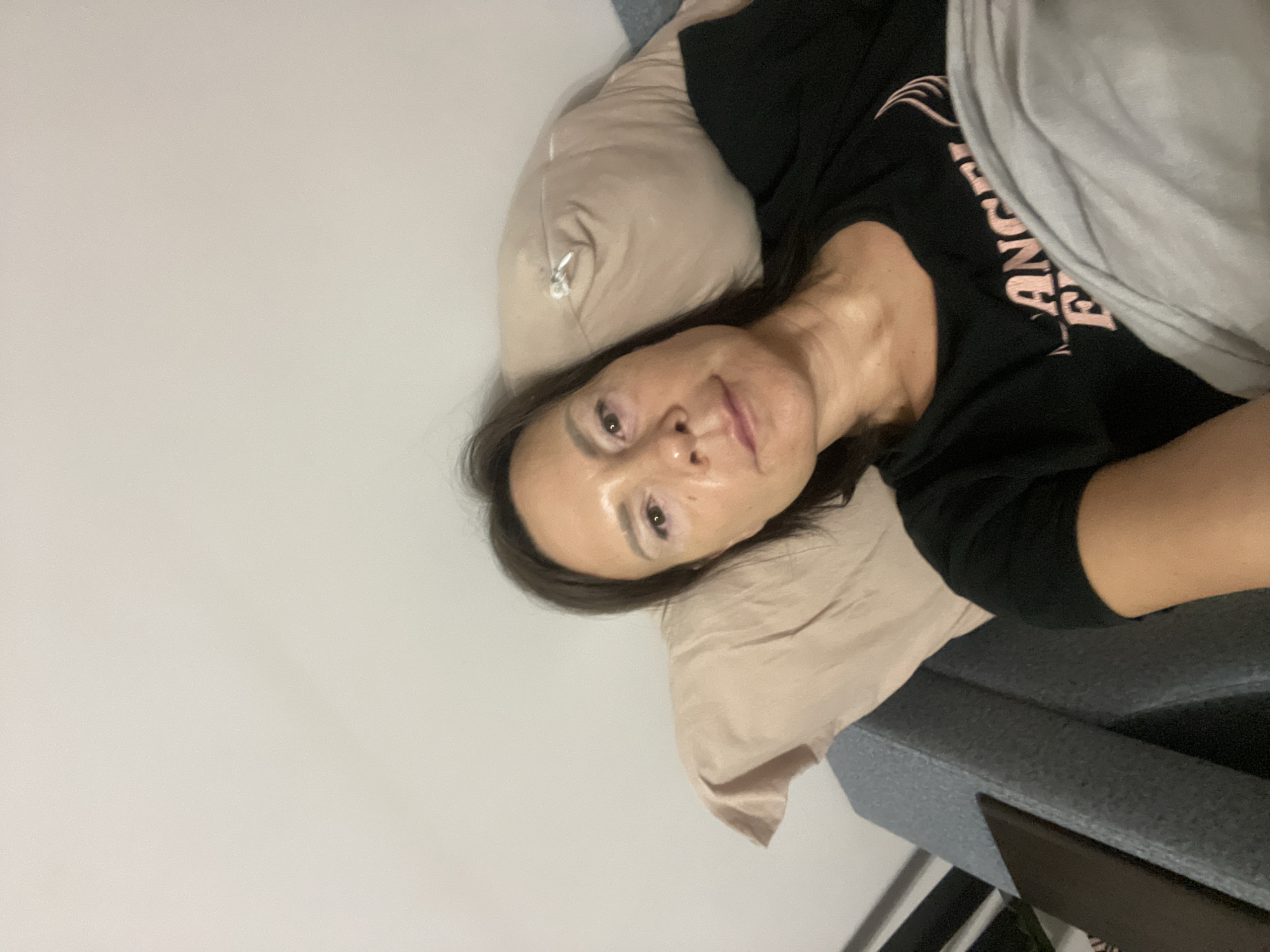 A person is lying in bed. The person has brown hair and is wearing a black shirt with pink lettering. A beige pillow supports the person's head. A gray blanket covers part of the person's body. The background shows a white wall and part of a gray upholstered bed frame. There is no information to place this image in Sochi, Russia.