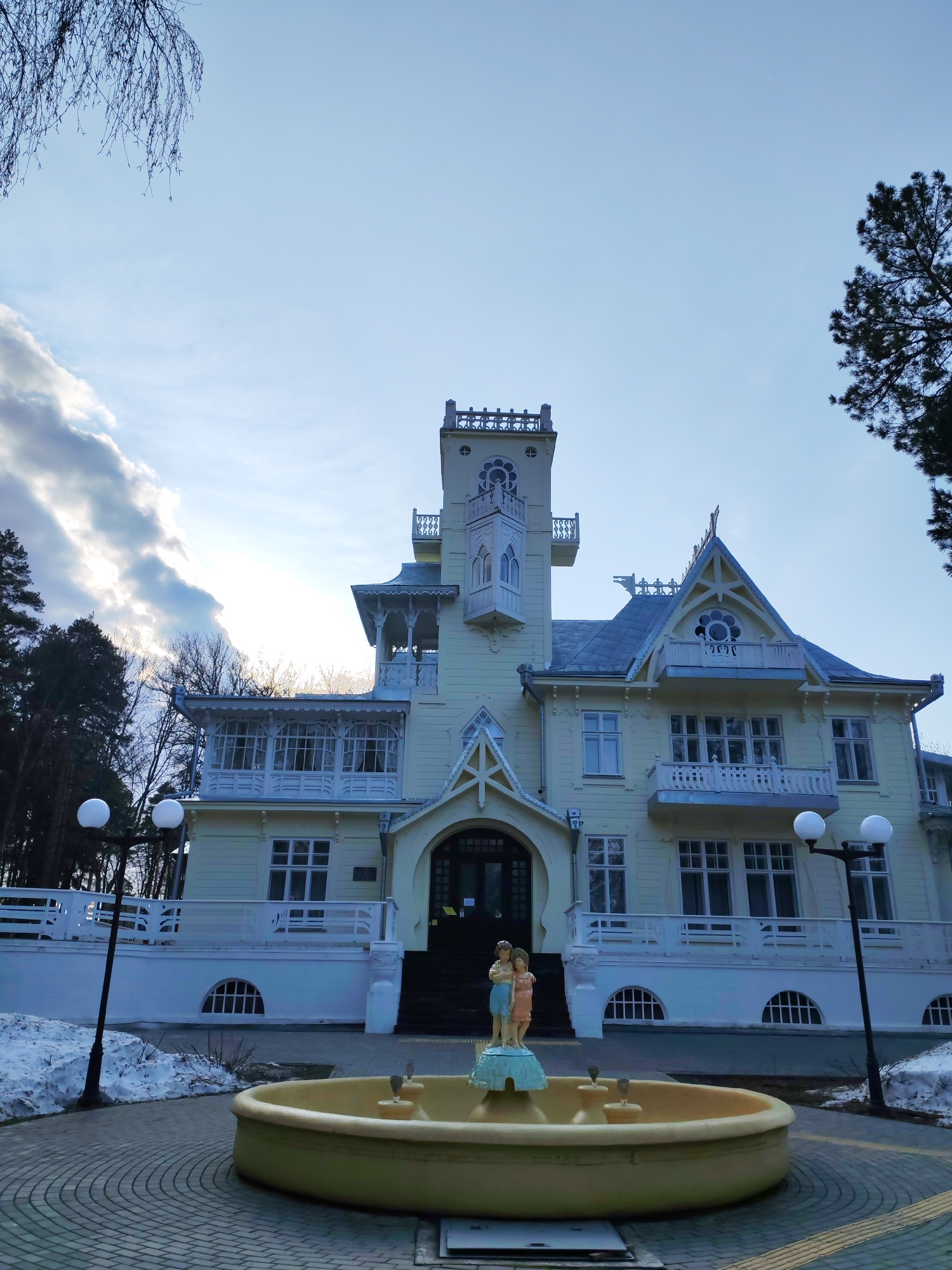 The image depicts a three-story building with a tower, located in Sarapul, Russia. The building is painted in light yellow, with white trim on the balconies and roof. A fountain in the foreground features a statue of two children. The surrounding area is paved with bricks, with snow patches visible. Two street lamps flank the fountain. Trees are visible in the background. The sky is partly cloudy with some sun.