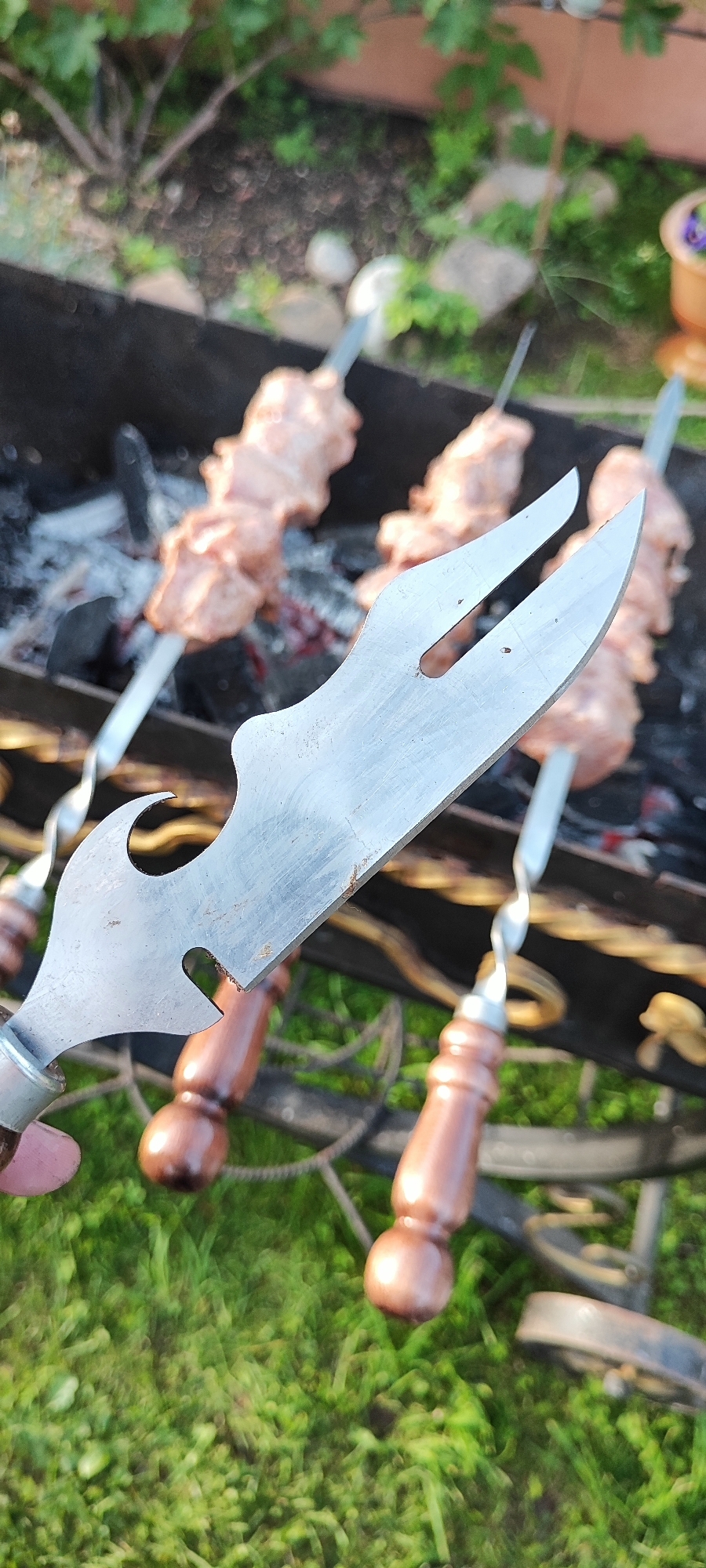 In an outdoor setting, possibly a backyard, a person is holding a metal skewer with raw meat on it, with a barbecue grill in the background. The skewer has a unique, curved metal handle with a bottle opener-like feature. The barbecue grill contains hot coals and several more skewers of meat. The ground is covered in green grass. The image appears to be taken during daylight. There are no visible landmarks or indicators to suggest the location is specifically in Лёзненскі сельскі Савет, Belarus.