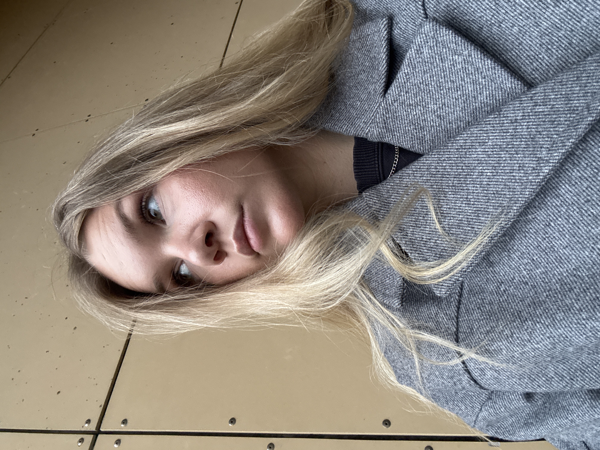 A young woman with blonde hair is lying on a surface that appears to be composed of rectangular panels, possibly a floor or a table. She is wearing a grey, herringbone-patterned coat. A black garment, possibly a top, is visible beneath the coat, and a delicate silver chain necklace is around her neck. The panelled surface beneath her is a light brown or tan color, marked with black lines and small metal rivets. The overall scene has an informal, candid quality. The location in Perm, Russia is speculative and can't be confirmed.