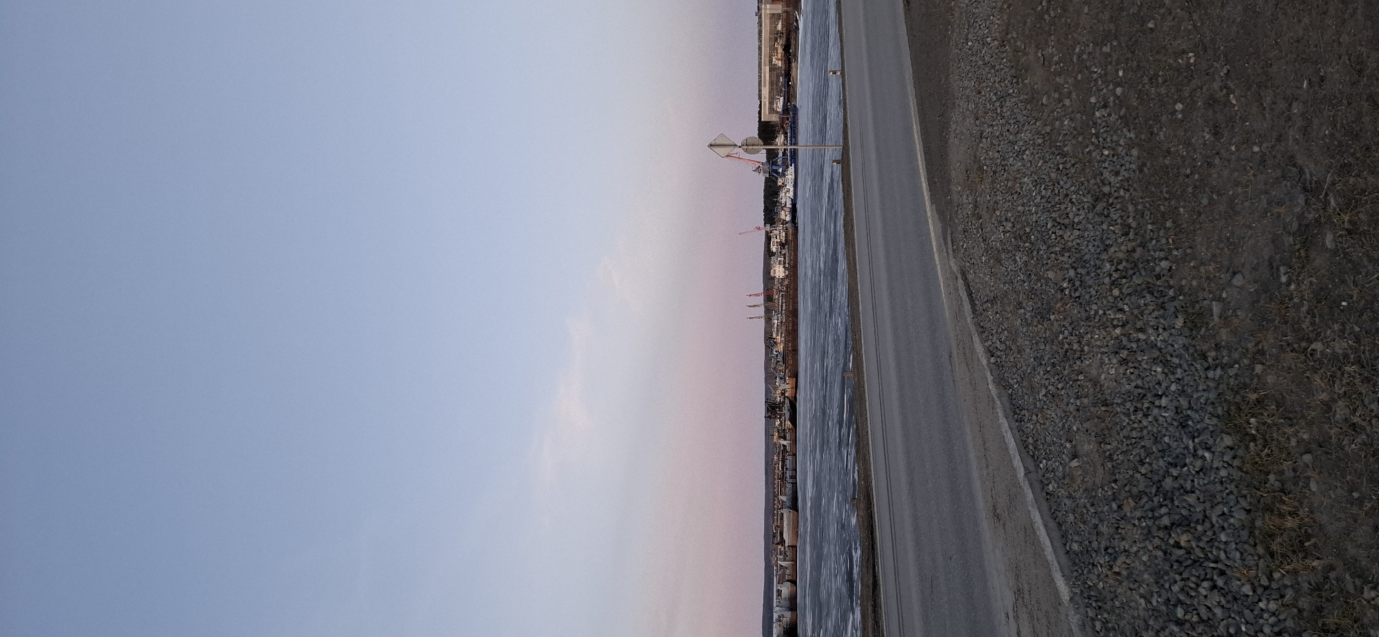 The image shows a view of the city of Chaikovsky, Russia at dusk or dawn. A body of water, likely the Kama River, is visible adjacent to the city's shoreline. The city's skyline consists of buildings, some of which are under construction with cranes present. A road with a guardrail runs parallel to the water's edge. The foreground consists of a gravelly or rocky area. The sky is a gradient of light blue to pink, indicating the time of day. A road sign is visible along the road. The city appears to be moderately industrialized.