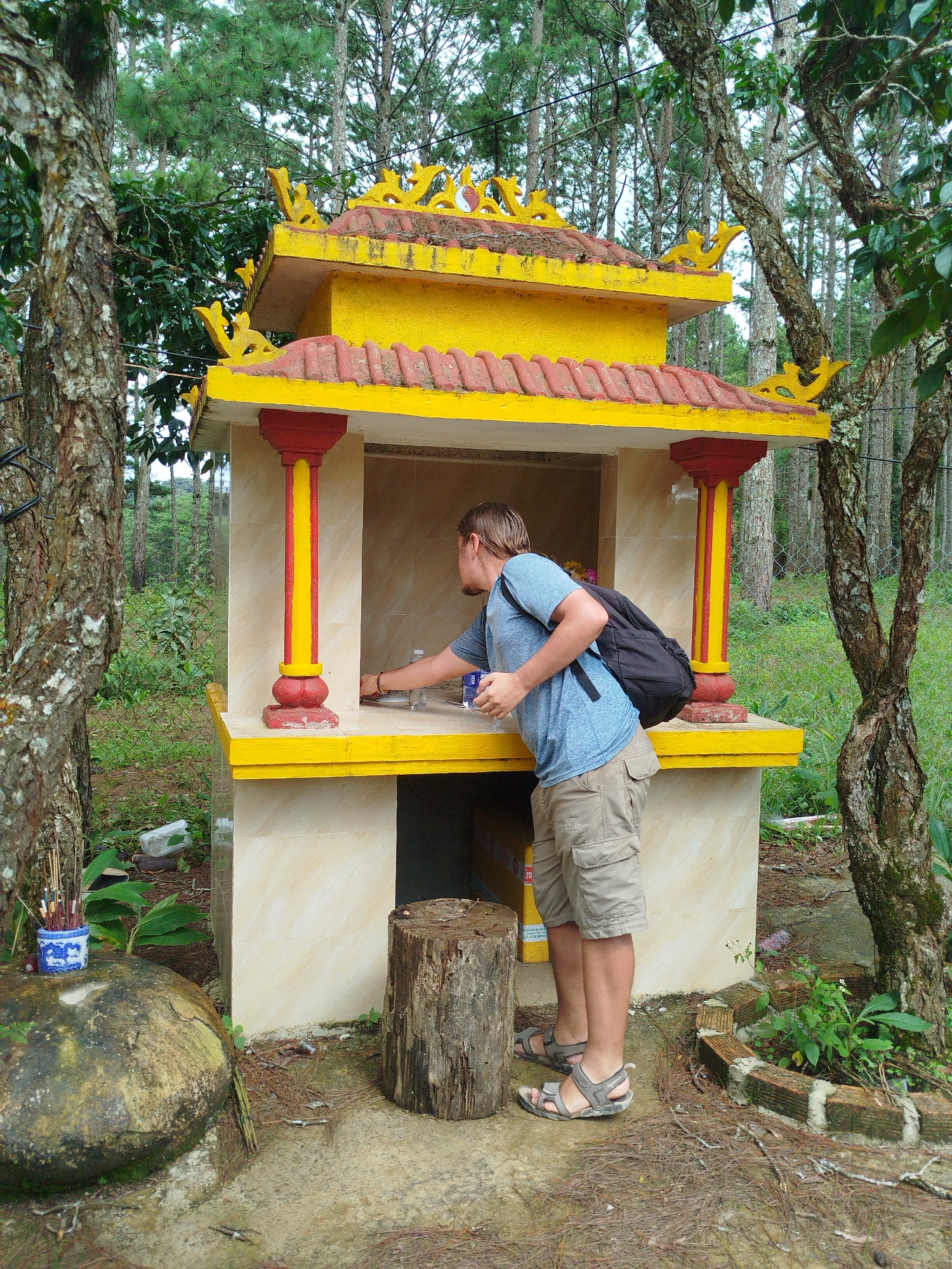 In Trạm Hành 1, Vietnam, a young man stands before a small, yellow and red shrine in a wooded area. He wears a blue t-shirt, beige cargo shorts, and sandals, with a black backpack slung over his left shoulder. He is reaching into the shrine, possibly to place or retrieve an item. The shrine is adorned with decorative yellow trim and has red and yellow pillars. In front of the shrine, a weathered log serves as a makeshift stool, and a large, moss-covered rock sits to the left. Incense sticks in a blue and white container are placed on the rock. The background consists of dense trees with thin trunks, suggesting a forest or park setting. The ground is covered with pine needles and scattered leaves, with a wire fence visible in the middle ground.