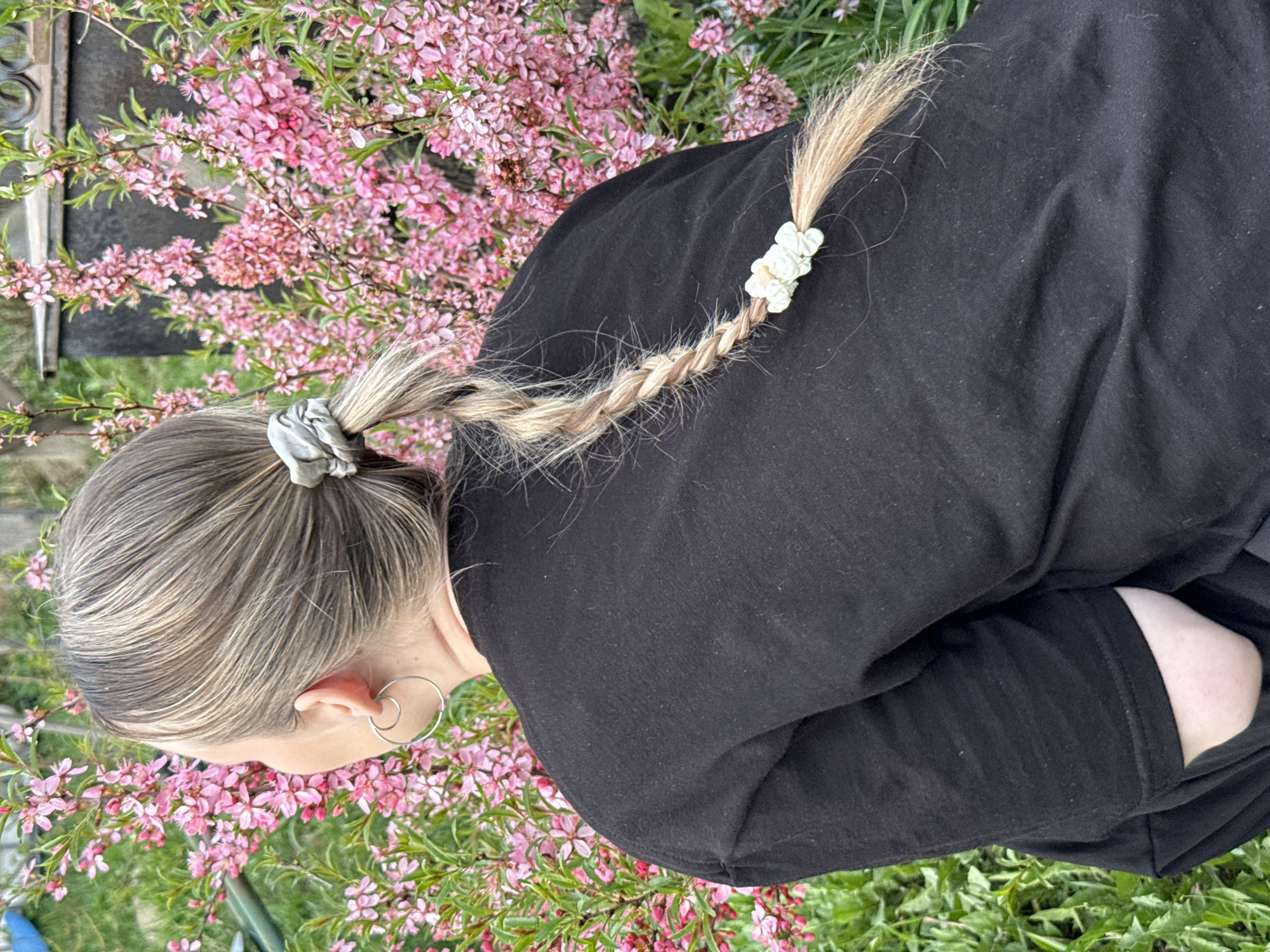 A high-angle outdoor shot shows a person with light brown and blonde hair standing near a flowering bush. The person wears a black long-sleeved shirt and has their arms crossed, obscuring their torso. Their hair is styled in a braid, secured with a scrunchie at the base and a white floral-patterned hair tie further down the braid. A silver hoop earring is visible on their left ear. The flowering bush features numerous small pink blossoms against green foliage. The location is identified as Unknown, Russia. Green grass is visible at the bottom of the frame.