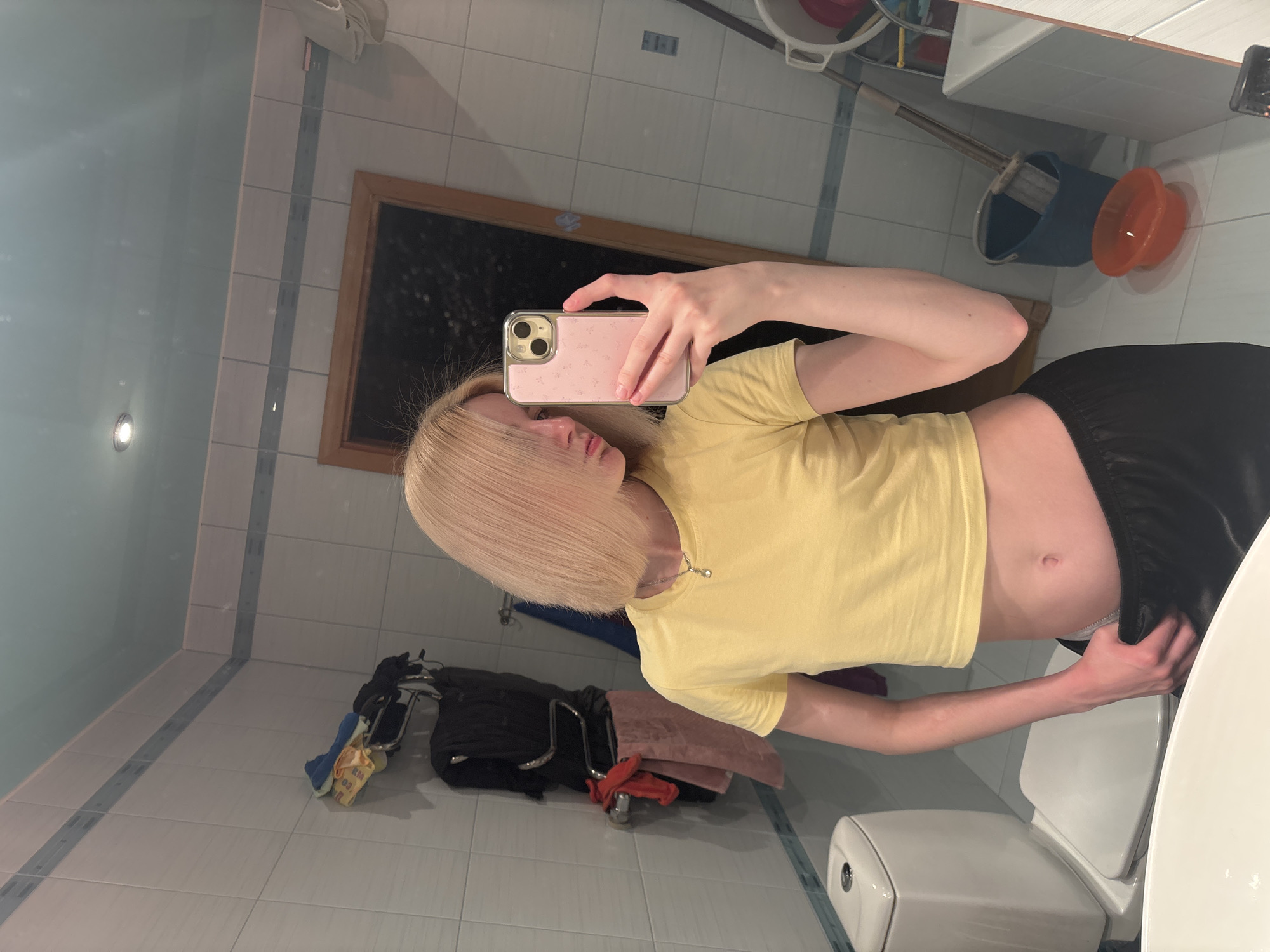 The image shows a young woman with blonde bob-cut hair taking a mirror selfie in a bathroom. She is wearing a yellow crop top and black pajama bottoms. The bathroom has tiled walls and a mirror above a sink. In the background, there is a toilet, a towel rack with towels, and cleaning supplies including a bucket and mop. The location is specified as Sinyavino, Russia.