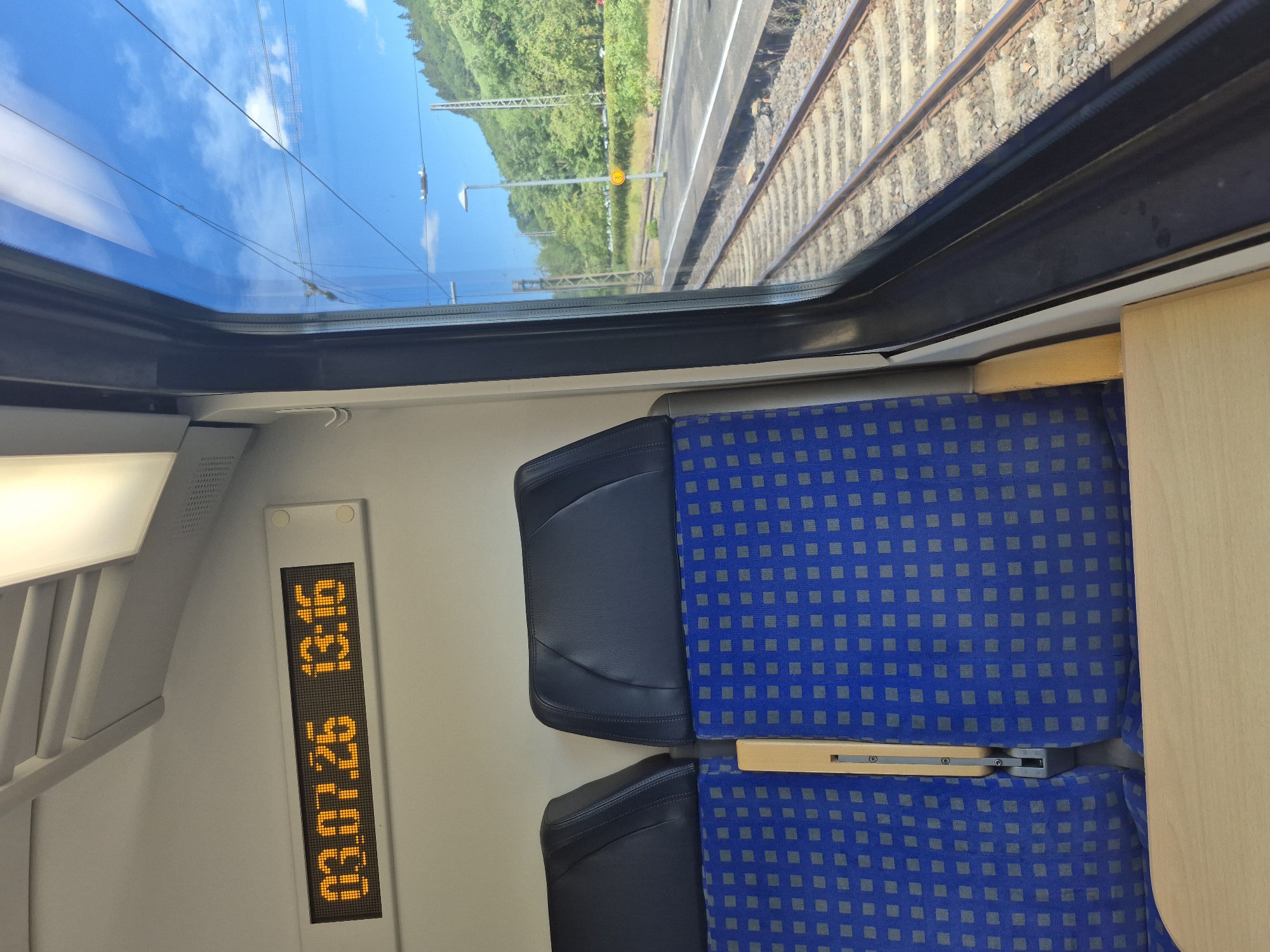 The image shows the interior of a train carriage with two blue and grey patterned seats visible. To the left of the seats, an orange digital display shows the date "03.07.26" and the time "13:16". Through the window on the right, railway tracks are visible, alongside a platform and a hillside covered in trees. The sky is blue with some white clouds. The train appears to be stopped at a station in Kobern-Gondorf, Germany, as suggested by the presence of the station and the surrounding landscape.