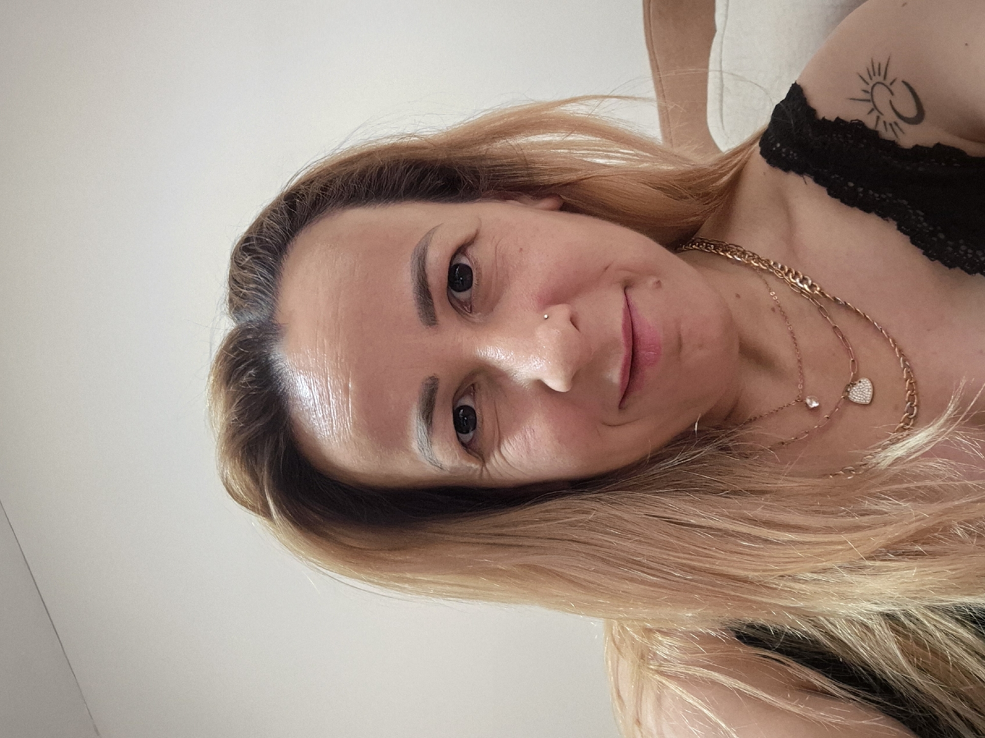 A woman with blonde hair and fair skin takes a selfie. She is wearing a black top with lace trim and several necklaces, one with a heart-shaped pendant. She has a nose ring and a tattoo of a sun and moon on her left shoulder. She is smiling slightly and looking at the camera. The background is a plain, light-colored wall and a blurred couch. The lighting is bright, and the focus is on the woman's face.