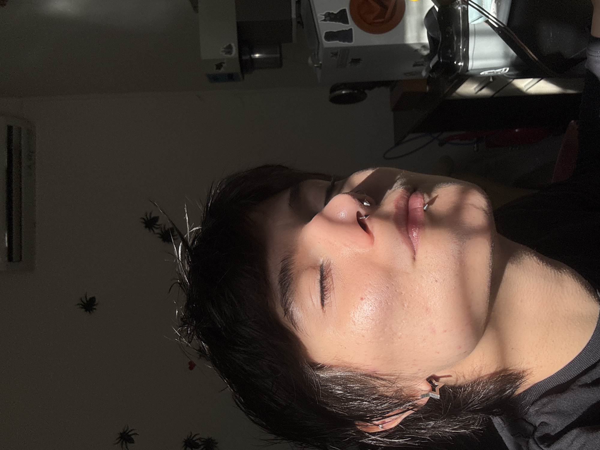 This indoor portrait, taken in Sofia, Bulgaria, features a young person resting with their eyes closed, captured from a side angle. Strong, directional sunlight illuminates their face, creating a striking contrast between brightly lit areas and deep shadows. The light highlights their features, particularly their nose and lips.
The person has dark hair and wears several piercings: a metallic ring through their septum and a barbell-style piercing below their lower lip. A silver-colored, star-shaped earring is visible in their left ear. Their closed eyes convey a sense of peacefulness, contemplation, or relaxation.
The background elements are largely in shadow. On the wall to the upper left, several black, decorative spider cut-outs are affixed, along with a white air vent or grille. In the upper right corner, above the person's head, parts of a shelf or desk are visible, holding indistinct objects including a round, dark orange item and other miscellaneous pieces. The strong sunlight suggests the photo was taken during either early morning or late afternoon, as the sun streamed through a window. There is no visible text in the image.