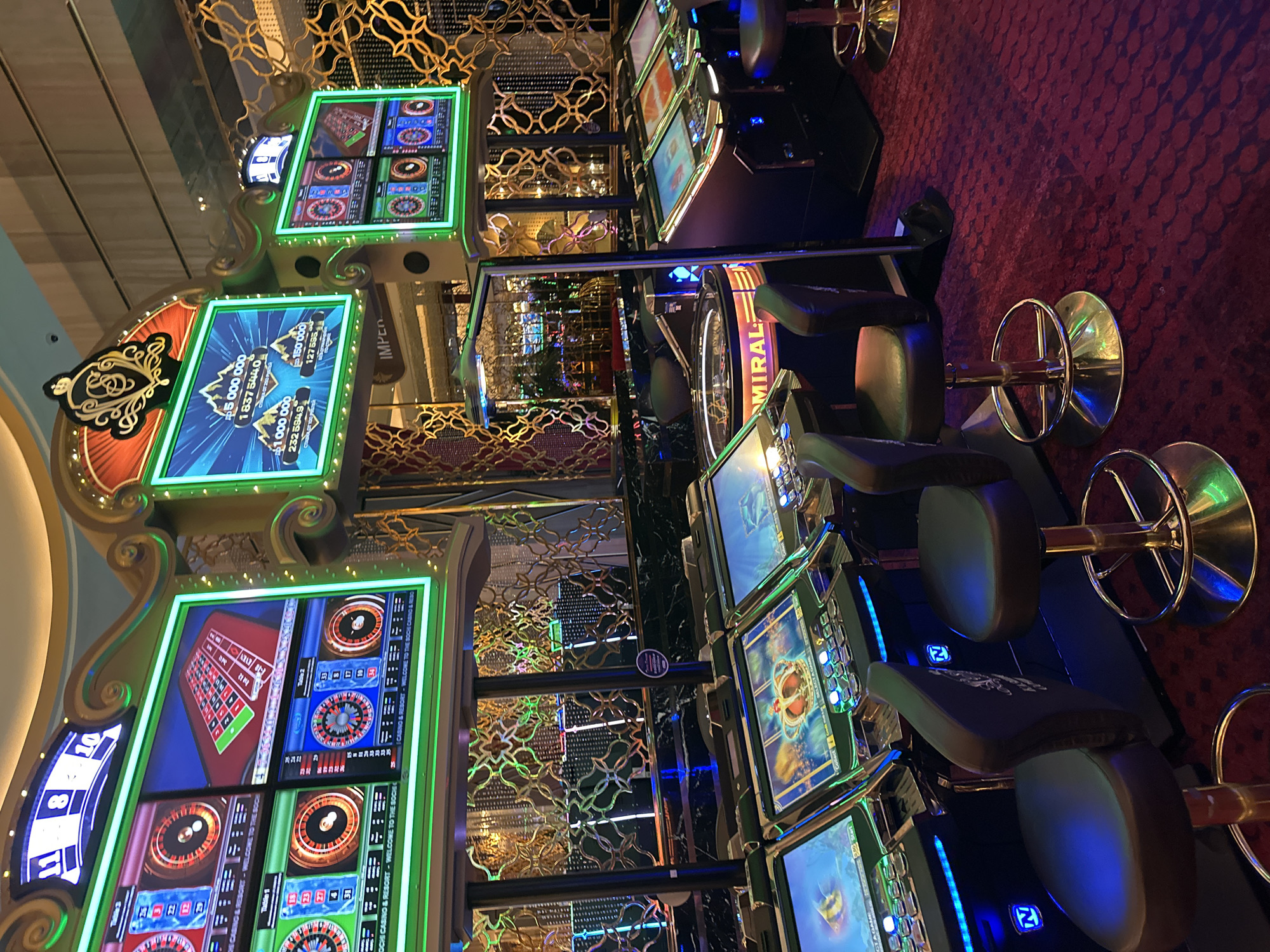 The image depicts an interior view of a casino, specifically within the Krasnaya Polyana resort in Estosadok, Russia, as indicated by the visible text "KASINO & RESORT - WELCOME TO THE SOCHI CASINO & RESORT". The scene is dominated by rows of electronic slot machines and roulette tables, bathed in the neon glow of various displays. Several casino chairs, upholstered in dark leather with gold-colored bases, are positioned in front of the machines. The floor is covered in a plush red carpet with a repeating dark pattern. In the background, elaborate golden decorative elements and lighting fixtures suggest a luxurious and opulent environment. One of the larger screens displays animated graphics and large numerical figures, likely indicating jackpots or prize amounts. The overall atmosphere is one of a modern entertainment establishment. There are no people clearly visible in the frame.