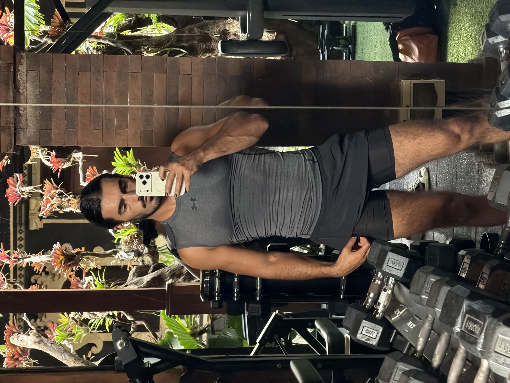 A man with dark hair and a mustache is taking a selfie in a gym mirror. He is wearing a grey sleeveless athletic shirt and black shorts. In the background, there are gym equipment, plants, and a brick wall. The gym appears to be outdoors or in a tropical setting, with lush greenery and ornate wooden structures. The overall lighting suggests it is daytime. The man is holding a white smartphone in his right hand.