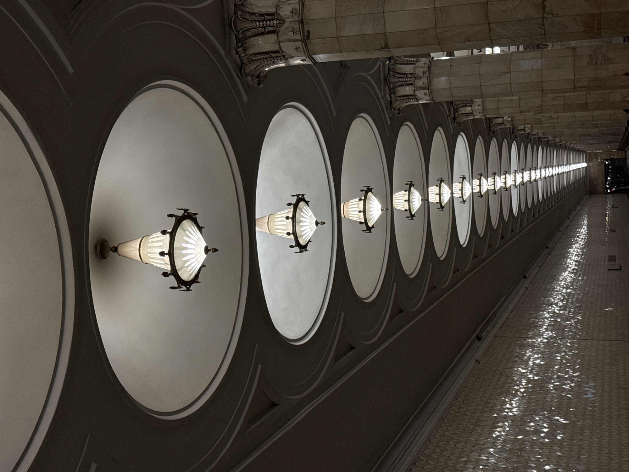 The image depicts a subway station interior in Moscow, Russia. A row of illuminated, conical-shaped lamps are recessed within circular openings along the ceiling of the station. The light fixtures have dark metal detailing. These openings are framed by dark gray borders. Columns with detailed ornamentation support the ceiling. The floor is paved with small, rectangular tiles that reflect the light, creating a glimmering effect. In the distance, the corridor narrows and fades into darkness. A dark vehicle is visible at the very end of the passage. No people are visible in the foreground.