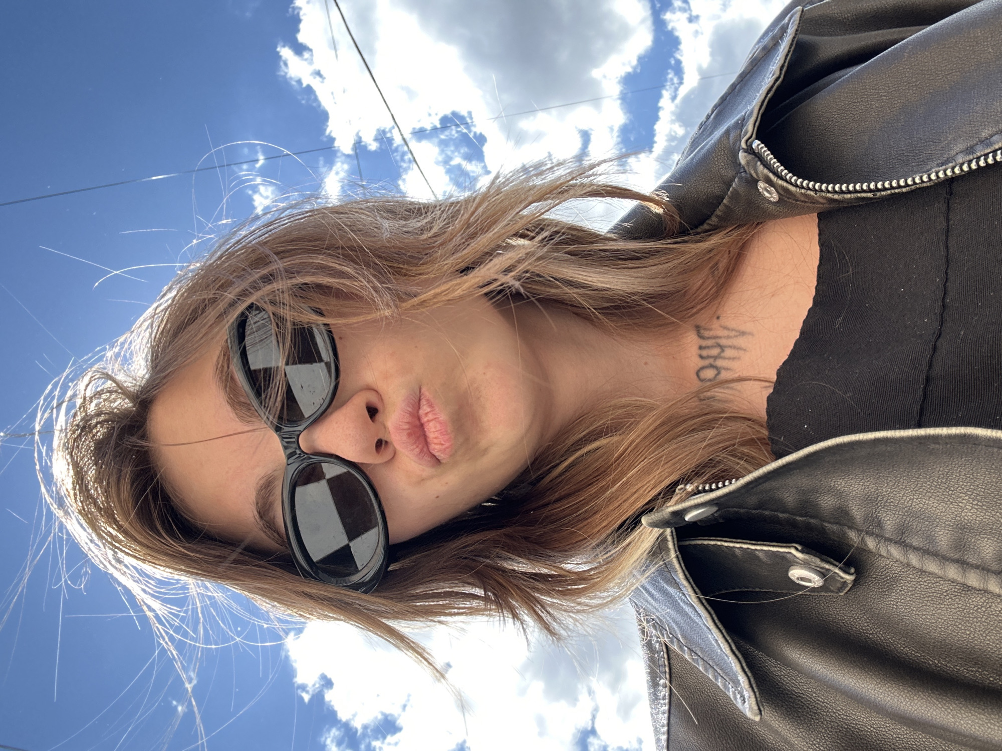 A low-angle, close-up, selfie-style photograph shows a young woman with blonde hair and sunglasses, making a kissing face. She is wearing a black leather jacket over a black top. She has a tattoo on her neck that reads "1998". The background consists of a bright blue sky with scattered white clouds and some telephone wires. The image does not provide enough information to determine the location in Moscow, Russia.