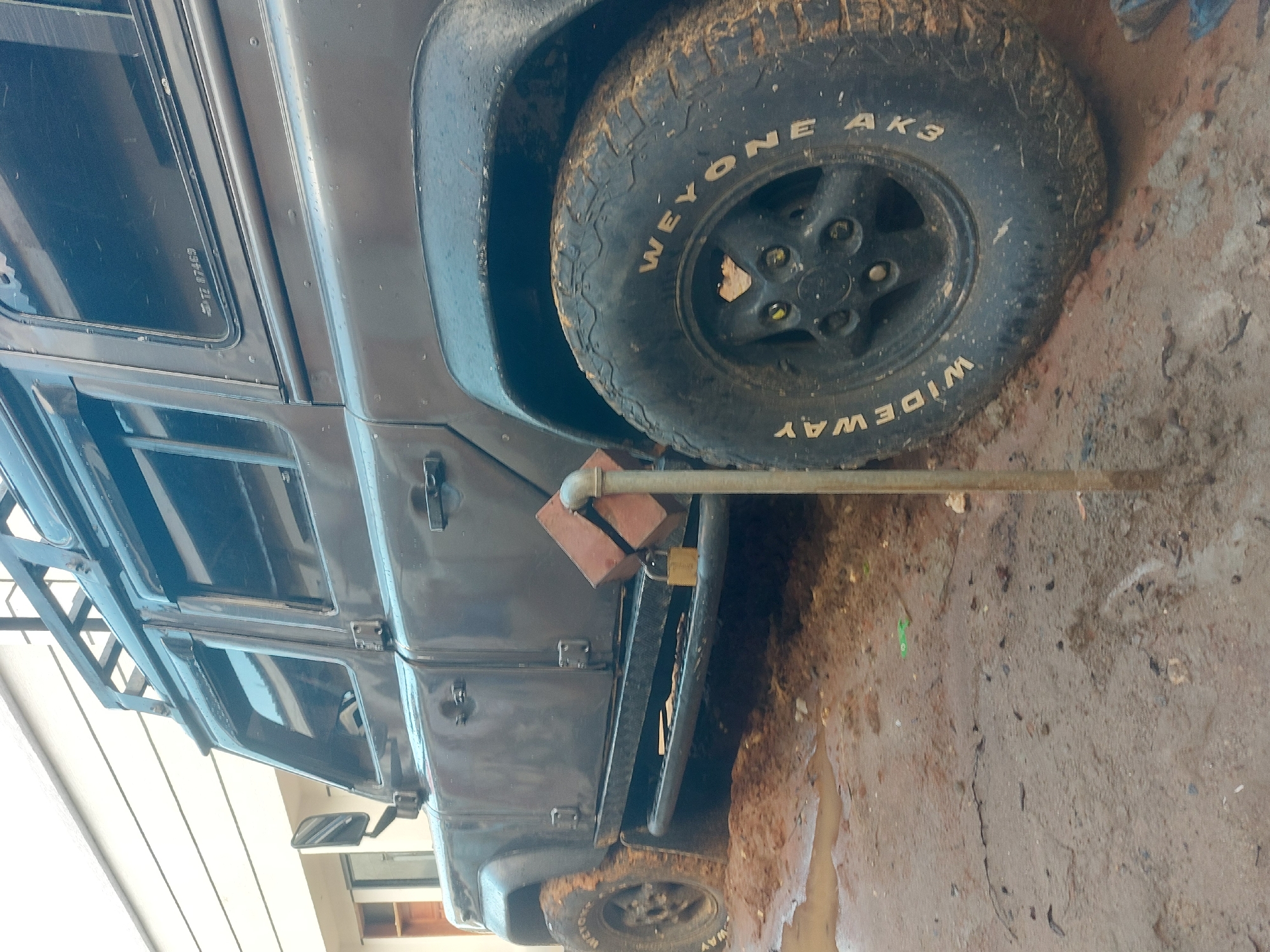 An overhead-angled shot captures the side of a dark-colored, rugged utility vehicle, likely a Land Rover Defender, heavily soiled with mud. The vehicle is stationary on a muddy, dirt ground, with puddles of water suggesting recent rainfall.
The main subject is the vehicle, which appears to be dark grey or black. Its large, knobby off-road tires are covered in mud, with white lettering visible on the sidewall of the prominent front right tire (from the viewer's perspective). The text "WEYONE AK3" and "WIDEWAY" can be clearly read. The vehicle features a robust roof rack and heavy-duty side steps. A metal pipe or bar is propped against the side step, secured by two red bricks and a brass padlock, an unusual setup that might serve as a temporary support or security measure.
On one of the rear side windows, there is visible text: "TZ 87469", which is likely a vehicle registration number, with "TZ" being the country code for Tanzania, aligning with the provided location context of Dar es-Salaam. The scene is illuminated by daylight, and no people are visible. The overall impression is one of a vehicle used in challenging, muddy conditions.