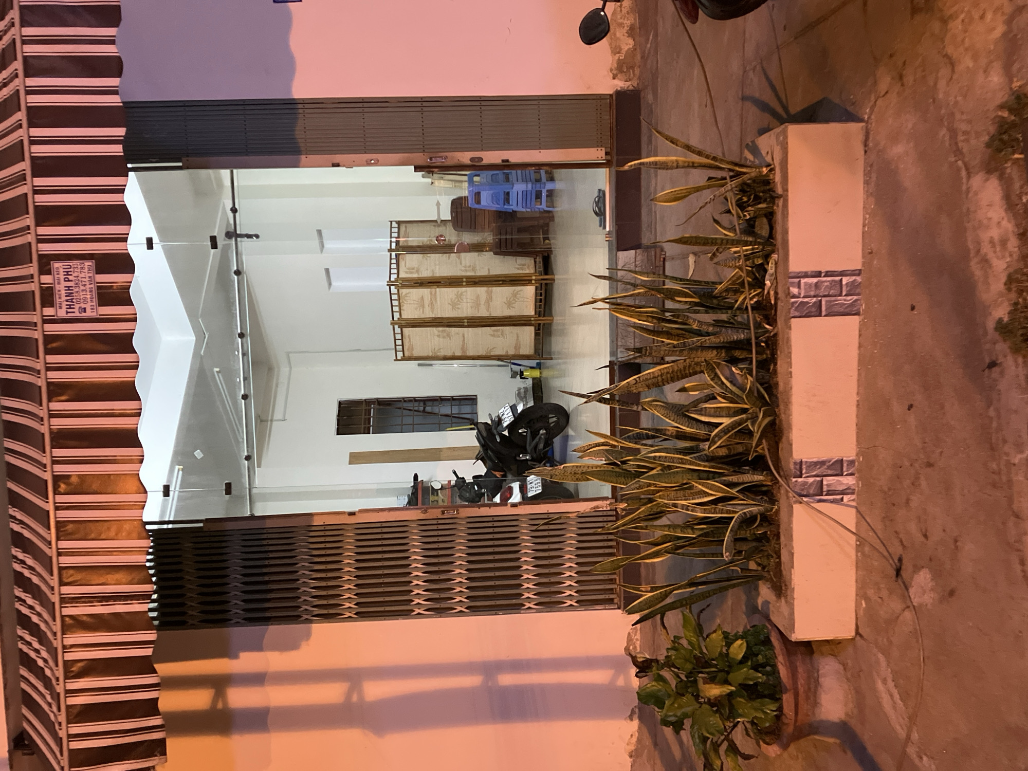 The image shows the entrance to a shop in Nha Trang, Vietnam, at night. The shop appears to be located on a street, with a striped awning over the entrance. The entrance itself is a large glass door, which is open, revealing the interior of the shop. Inside, there is a motorcycle parked, and a folding screen. In front of the shop, there are two planters with plants. The planter on the left is a round terracotta pot, and the planter on the right is a rectangular concrete planter. The ground in front of the shop is paved. There is a sign above the shop entrance that reads "THANH PHU" and lists a phone number.
