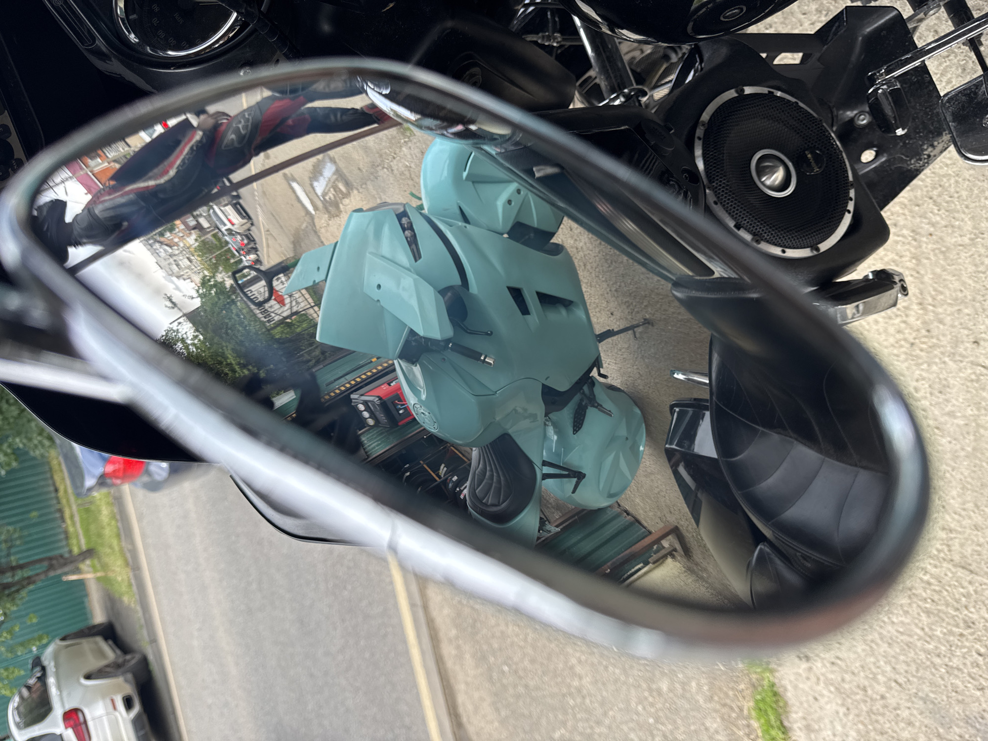 The image shows a reflection in a motorcycle mirror. The reflection reveals a light blue scooter parked on the street. A person wearing a black and red motorcycle suit is also reflected. In the background, there's a street scene with buildings and parked cars. The location appears to be Krasnodar, Russia. A portion of the motorcycle the mirror is attached to is visible, including a speaker.