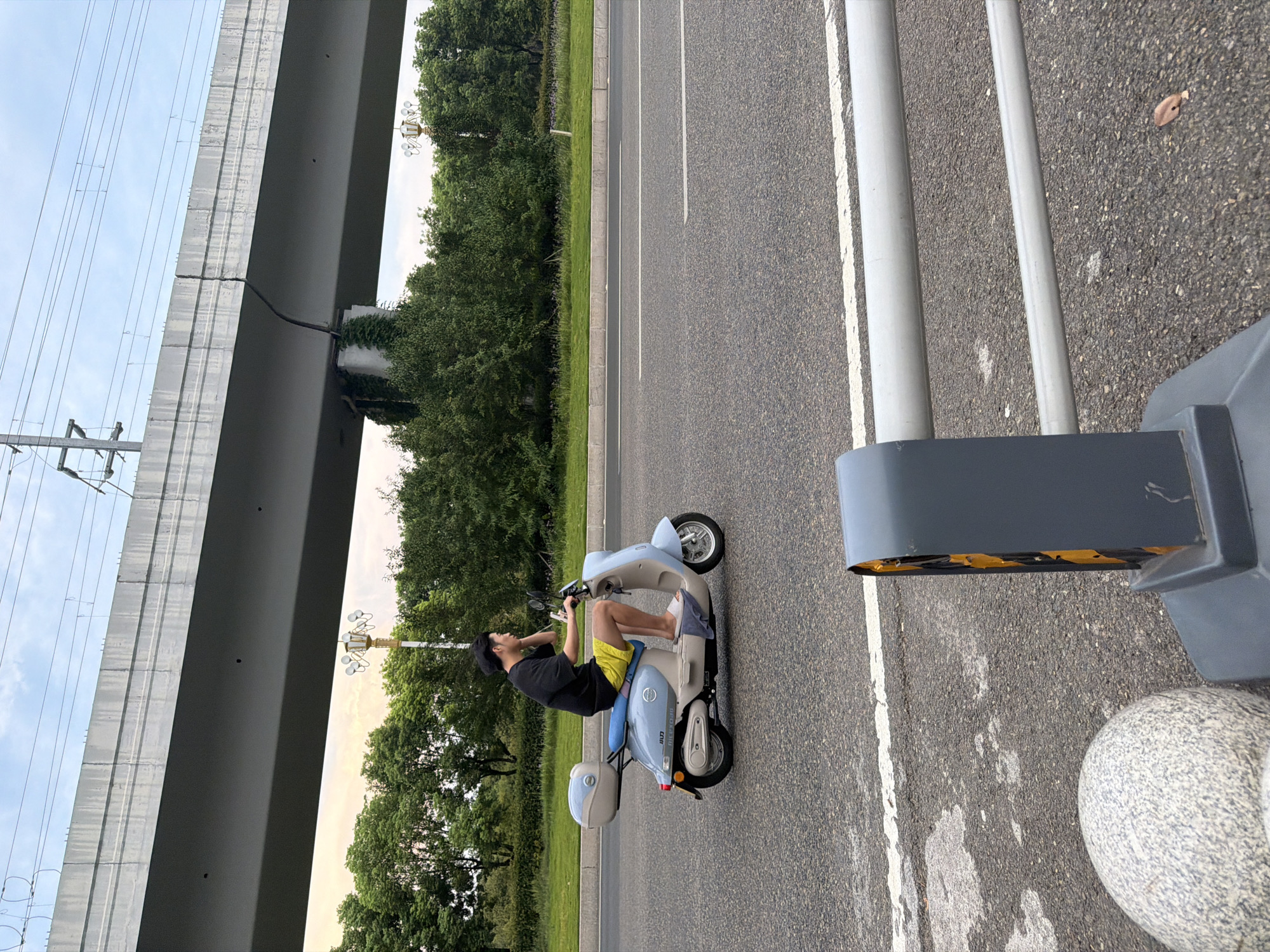 A man wearing a black t-shirt and yellow shorts rides a light blue electric scooter on a paved road. He has dark hair and is looking to his right. The scooter has a light blue storage box on the back and a blue seat. In the background, there is a large, elevated concrete structure, likely an overpass, with electrical wires running above it. To the left of the road is a grassy median with trees. The sky is overcast with some patches of lighter sky. In the foreground, there is a gray traffic barrier with reflective yellow markings and a rounded, grey stone object. The scene appears to be a road in an urban or suburban area, possibly in Jiangjin, China, given the presence of lush green trees and the style of the overpass.