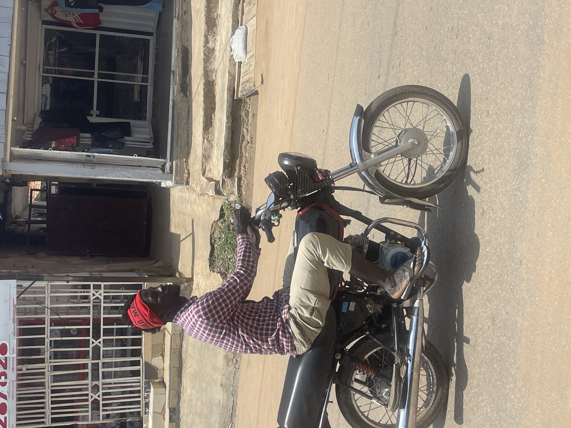 A man wearing a red and black checkered shirt, khaki pants, and a red bandana is riding a black motorcycle down a dusty, paved street. The motorcycle has a chrome exhaust pipe and a black headlight. Behind the man, a building with a white exterior and a metal grate entrance is visible. Further to the right, another building has a glass storefront with clothing displayed inside. The scene appears to be in an urban environment, likely a market area, given the shops. The lighting suggests it is daytime, with shadows cast by the motorcycle and the buildings. The ground is a mix of dirt and asphalt. There is no visible indication of this being in Abuja, Nigeria.