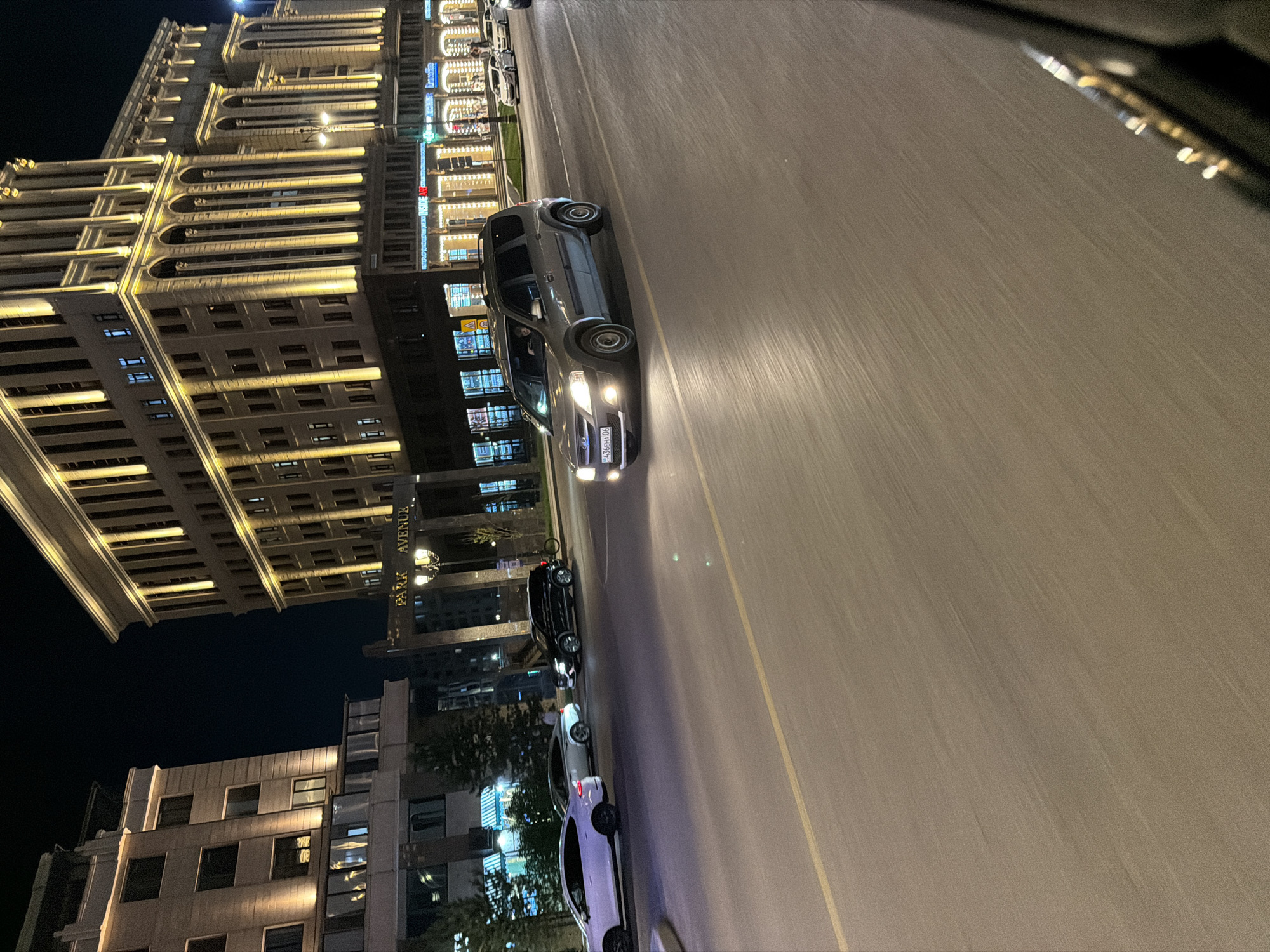 This is a night-time, eye-level, medium shot of a street in Astana, Kazakhstan, taken from inside a moving vehicle. The image features a wide asphalt road with a yellow line marking the lanes. Several cars are visible on the road, including a grey SUV in the foreground with its headlights on, and other cars further down the street, some parked and some moving.
Dominating the background are two large, illuminated buildings. The building on the right is a tall, multi-story structure with a classical facade, featuring numerous arched windows and vertical lighting elements. A sign above the entrance to this building reads "PARK AVENUE". The building on the left is also a multi-story structure, with a more modern architectural style, featuring horizontal window bands and some areas lit with warm yellow lights. Several trees are planted between the road and the buildings.
The overall scene depicts a busy urban street at night, likely a commercial or residential area in Astana, given the "Park Avenue" sign which is a known street name in the city. The motion blur indicates that the photograph was taken from a moving vehicle.