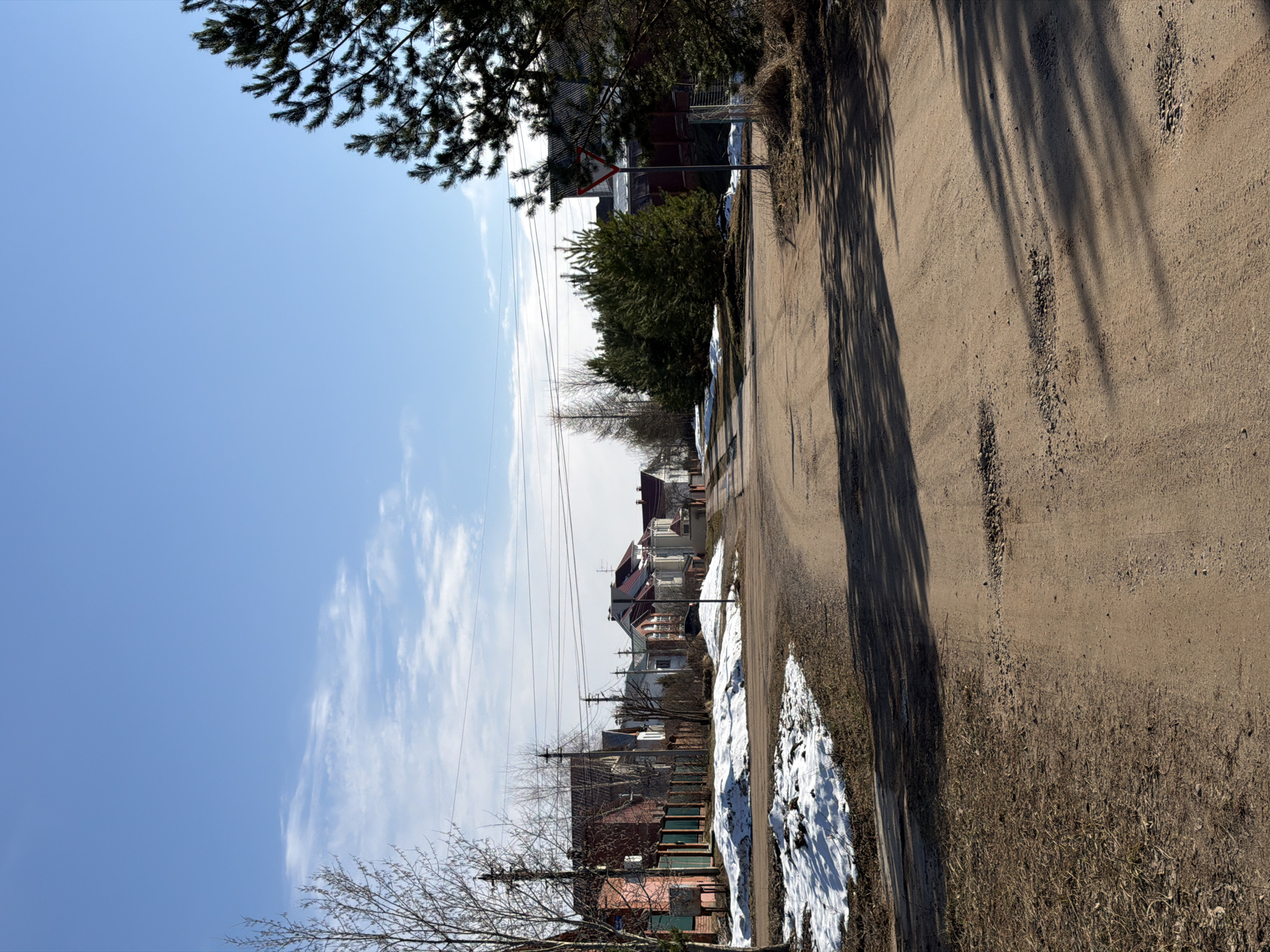 The image depicts a dirt road running through a residential area in Chaikovsky, Russia. The road is bordered by houses on the left side and vegetation on the right. Patches of melting snow are visible along the road's edge. Shadows cast by trees and buildings stretch across the road surface. A triangular road sign stands near the houses. Power lines traverse the sky above the houses. The sky is blue with scattered clouds. The angle of the image shows a street in Chaikovsky Russia.