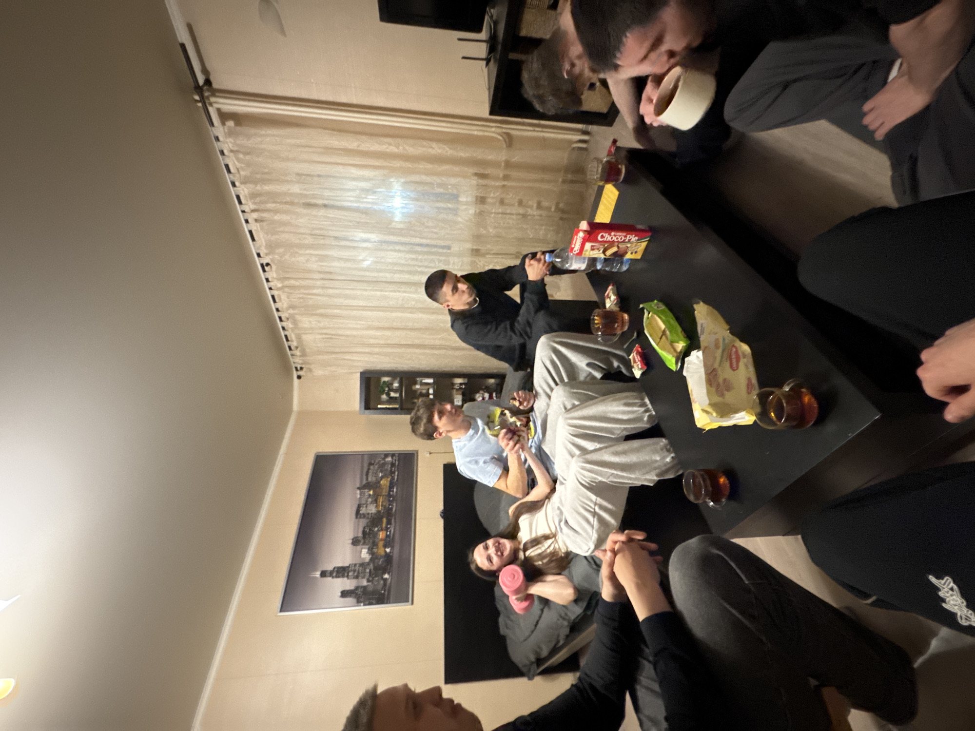 A group of young people are sitting around a black coffee table in an apartment. There are about seven people visible. Some are seated on a couch. On the table are snacks, drinks, and a box of Choco-Pie. In the background, there is a framed black and white cityscape on the wall. The location is likely Kazan, Russia, based on the cityscape potentially depicting the city skyline.