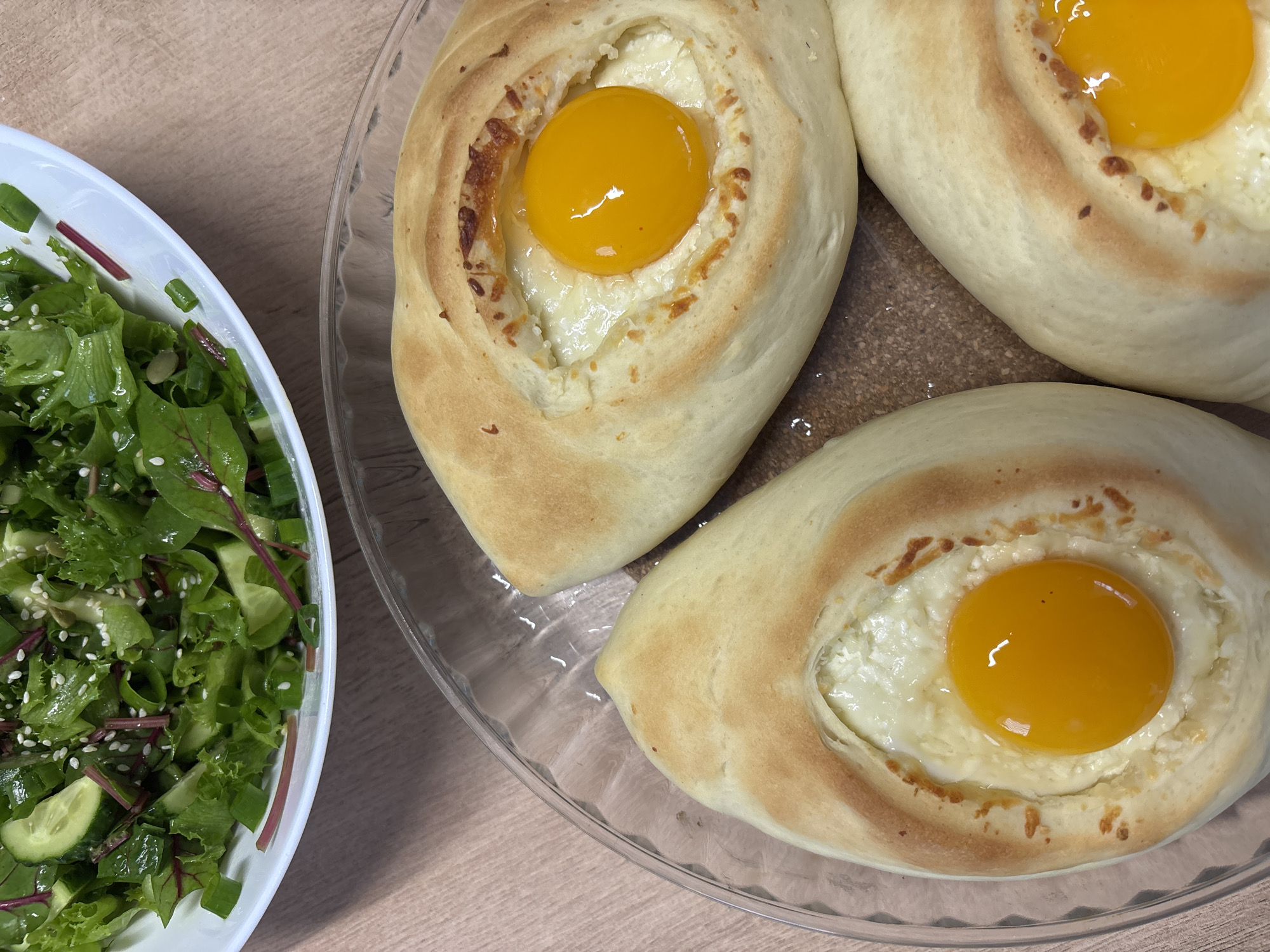 The image shows a top-down view of a meal consisting of three khachapuri adjaruli (Georgian cheese bread with egg) in a glass serving dish, placed next to a salad in a white bowl. The bread boats are golden brown and filled with cheese and a single egg yolk each. The salad contains a mix of green leaves, cucumber slices, green onions, and what appears to be sesame seeds. The meal sits on a light-brown wooden surface. The location of the photo is not discernible. The image does not contain people.