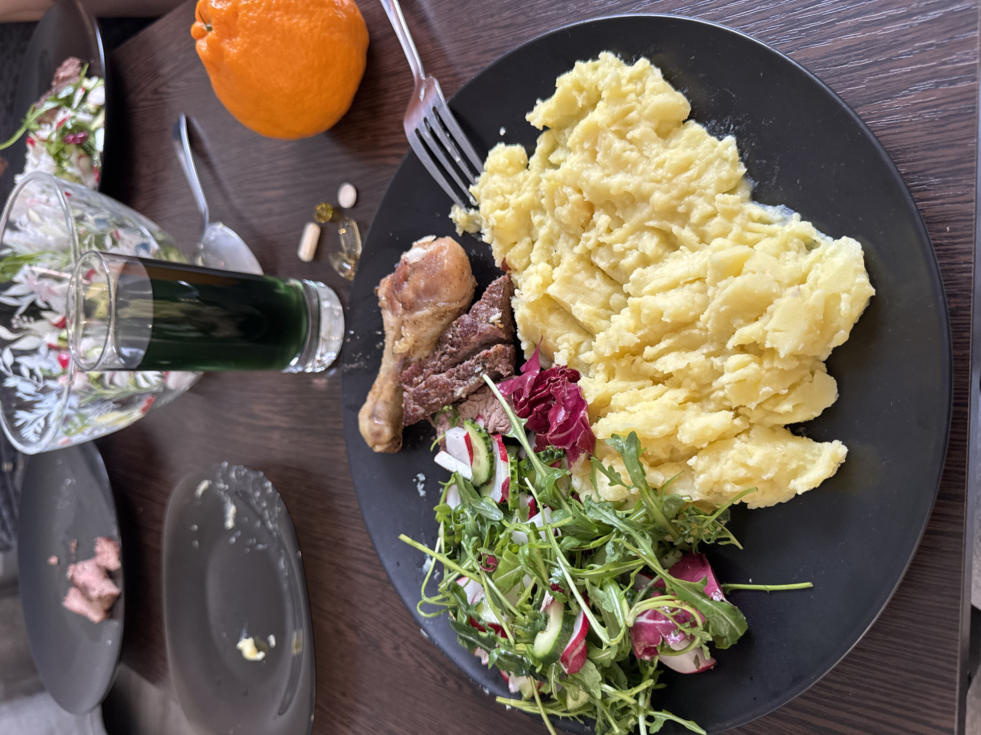 A meal is displayed on a dark brown wooden table, likely set indoors in Murino, Russia. The central feature is a large black, roundish plate holding a generous portion of light yellow mashed potatoes. Alongside the potatoes are a cooked chicken drumstick and several slices of what appears to be cooked red meat. A fresh salad, composed of green arugula, reddish radicchio leaves, sliced radishes, and cucumber, completes the main dish. A silver fork is resting on the plate, partially embedded in the mashed potatoes.
To the left of the main plate, a clear glass contains a dark green liquid, resembling a smoothie or juice. Further left, an orange fruit with a textured peel, possibly a tangelo or large clementine, is visible. Near the orange and the glass, a small collection of supplements or medications is arranged, including a white pill, a clear capsule with light contents, and two small yellow translucent capsules.
In the lower-left corner, two smaller dark gray plates are present; one is mostly empty with a few scattered crumbs, while the other holds a few small pieces of meat, suggesting that some parts of the meal may have been partially consumed or served. A silver spoon is partly visible beneath the orange and glass. The scene portrays a substantial and varied home-cooked meal. No people are visible, and there is no discernible text within the image. The lighting indicates it is likely daytime.