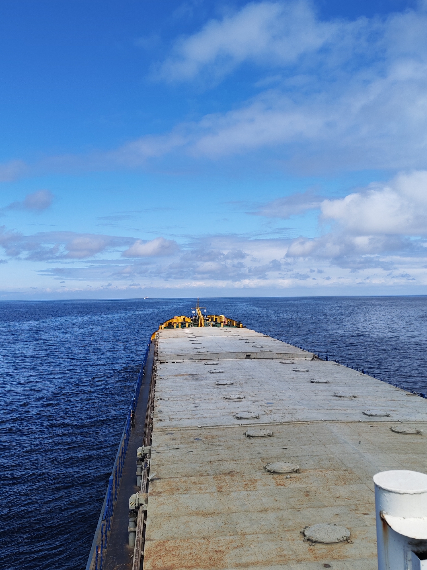 The image shows a view from the deck of a ship, likely a cargo vessel, sailing on a body of water, possibly the sea. The ship's deck takes up a significant portion of the foreground and appears to be made of large, flat, rectangular sections with several circular manhole covers. The deck shows signs of wear and tear, with rust and staining visible.
Beyond the ship, the sea stretches to the horizon. The water is a dark blue color, reflecting the sky. There are small waves on the surface. In the distance, a faint outline of another vessel can be seen.
The sky is predominantly blue, with scattered clouds of varying sizes and shapes. The clouds are mostly white, with some gray shading.
The exact location is unknown and there is no visual evidence to suggest the city is Unknown, Russia.