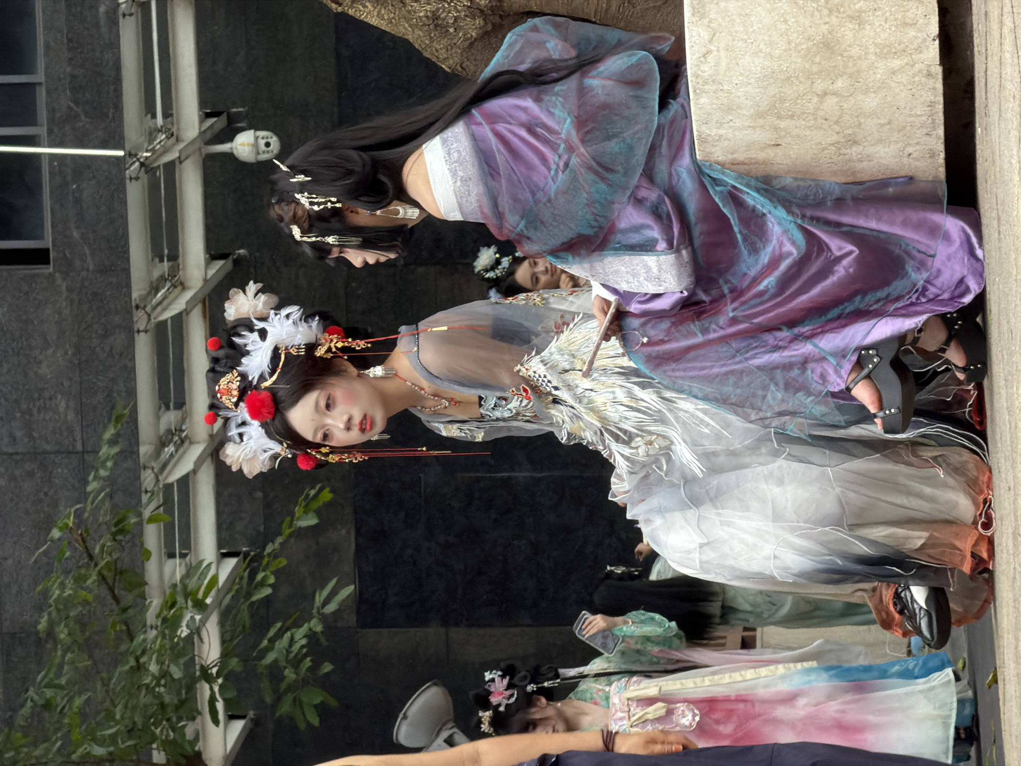 In Yuzhong District, China, two young women dressed in elaborate, traditional Hanfu-style clothing are the central focus of the image. The woman on the left, seated, wears a white and grey gradient robe adorned with intricate embroidery, featuring a feathered design. She has a detailed headdress with red pom-poms and white feathers, and her makeup is dramatic with accentuated red eyeliner. She holds a fan. The woman on the right is also seated and wears a flowing purple and teal iridescent robe, draped over her shoulders. Her hair is styled in an updo with delicate gold accessories. A third woman, partially visible on the far left, is wearing a pink and blue gradient dress and black sneakers. Behind them, a security camera is mounted on a white structure against a dark, textured building facade. Trees with green leaves are visible in the upper left corner. The women are seated on a light-colored, textured block.